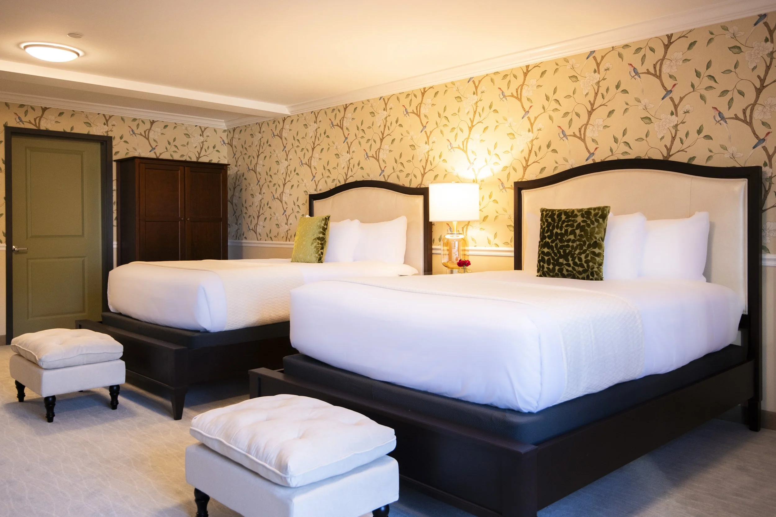 Two hotel beds with white bedding, green accent pillows, and a nightstand with a lamp in a decorated room with floral wallpaper and a wooden wardrobe.