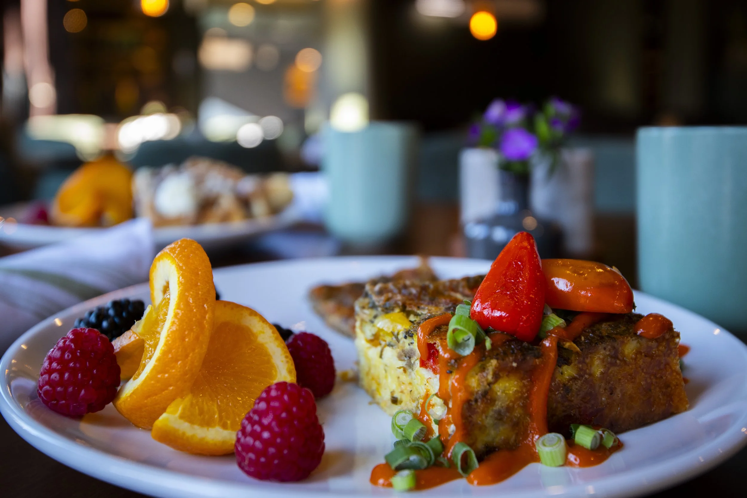A plate of breakfast food with orange slices, raspberries, blackberries, and a piece of egg casserole topped with cherry tomatoes, green onions, and red sauce, on a table in a restaurant setting.