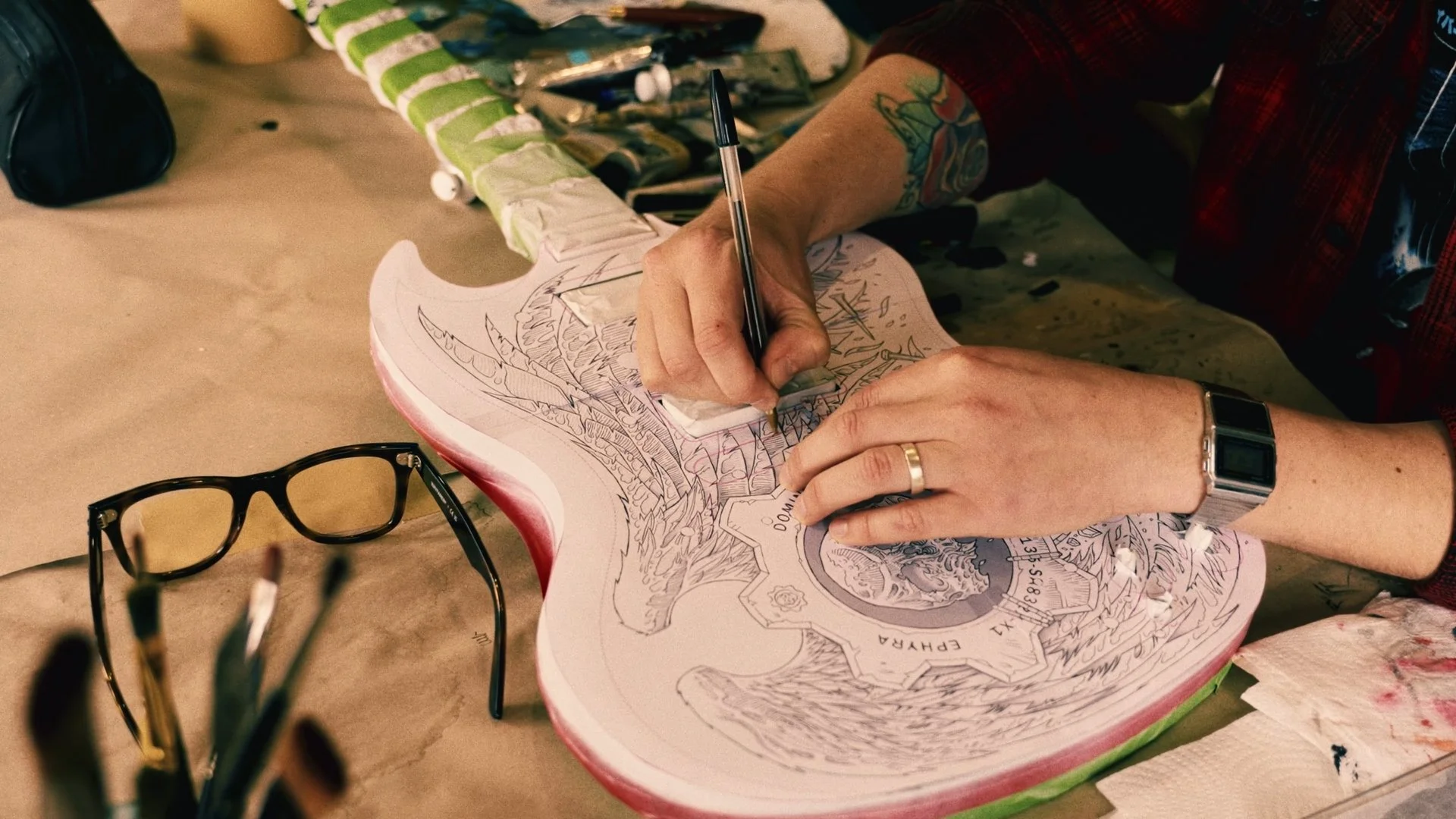 Person sketching on an artist's guitar with various art supplies and glasses nearby.