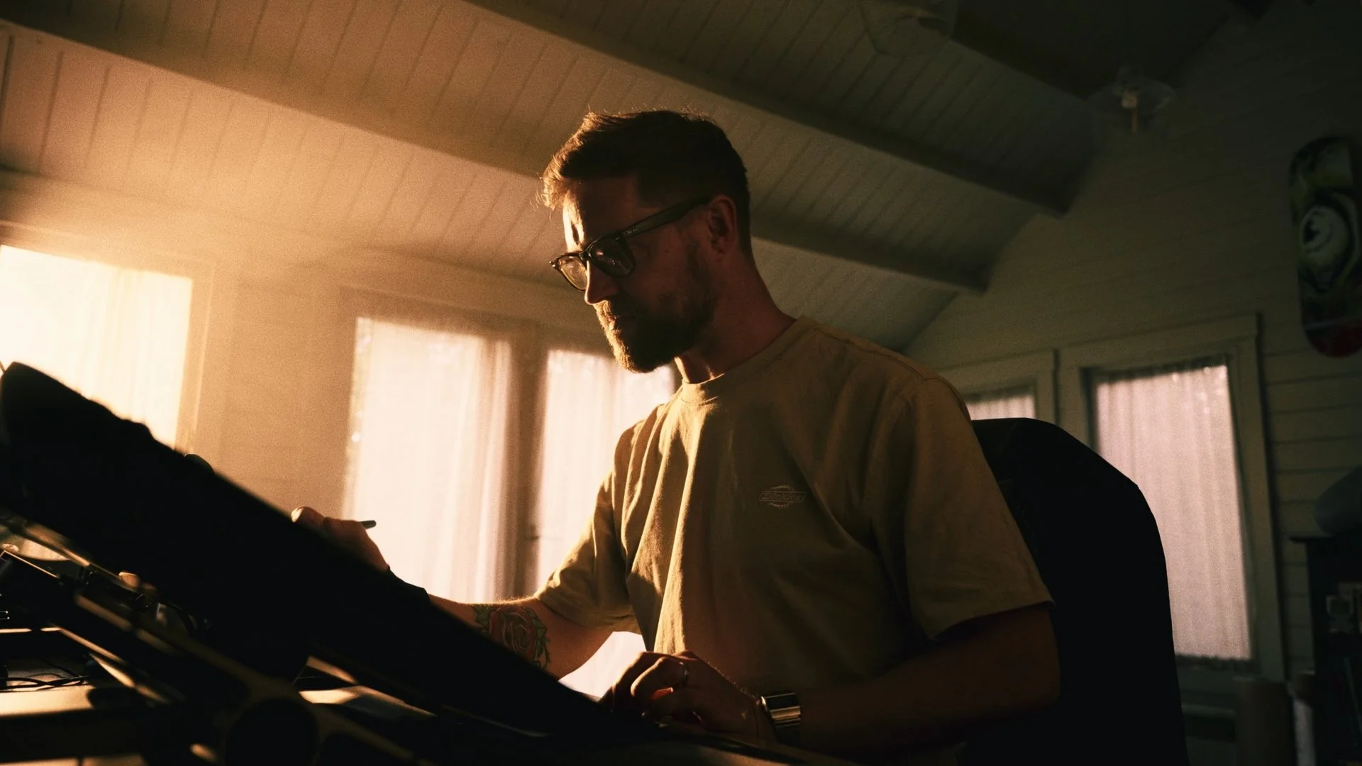 A man with glasses and a beard wearing a yellow T-shirt is sitting at a keyboard or some electronic music equipment in a dimly lit room with warm sunlight coming through the windows.