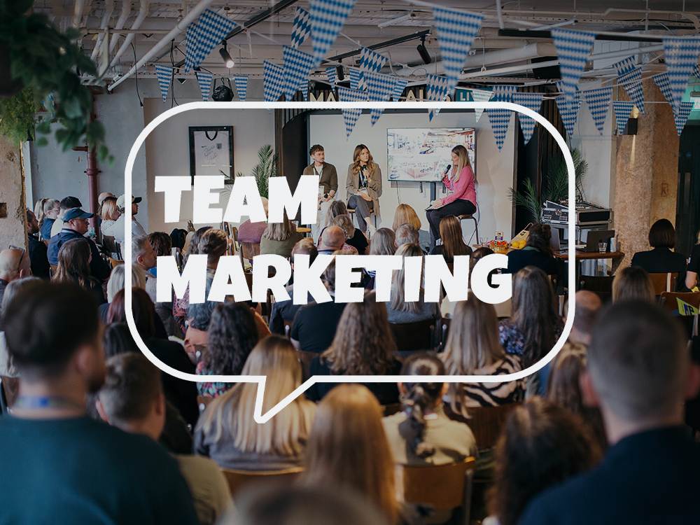 A large audience sitting and listening to a panel discussion at a marketing event. There are banners with blue and white checkered patterns hanging from the ceiling, and a speaker presenting on a stage with a large screen behind them. The text 'TEAM 
