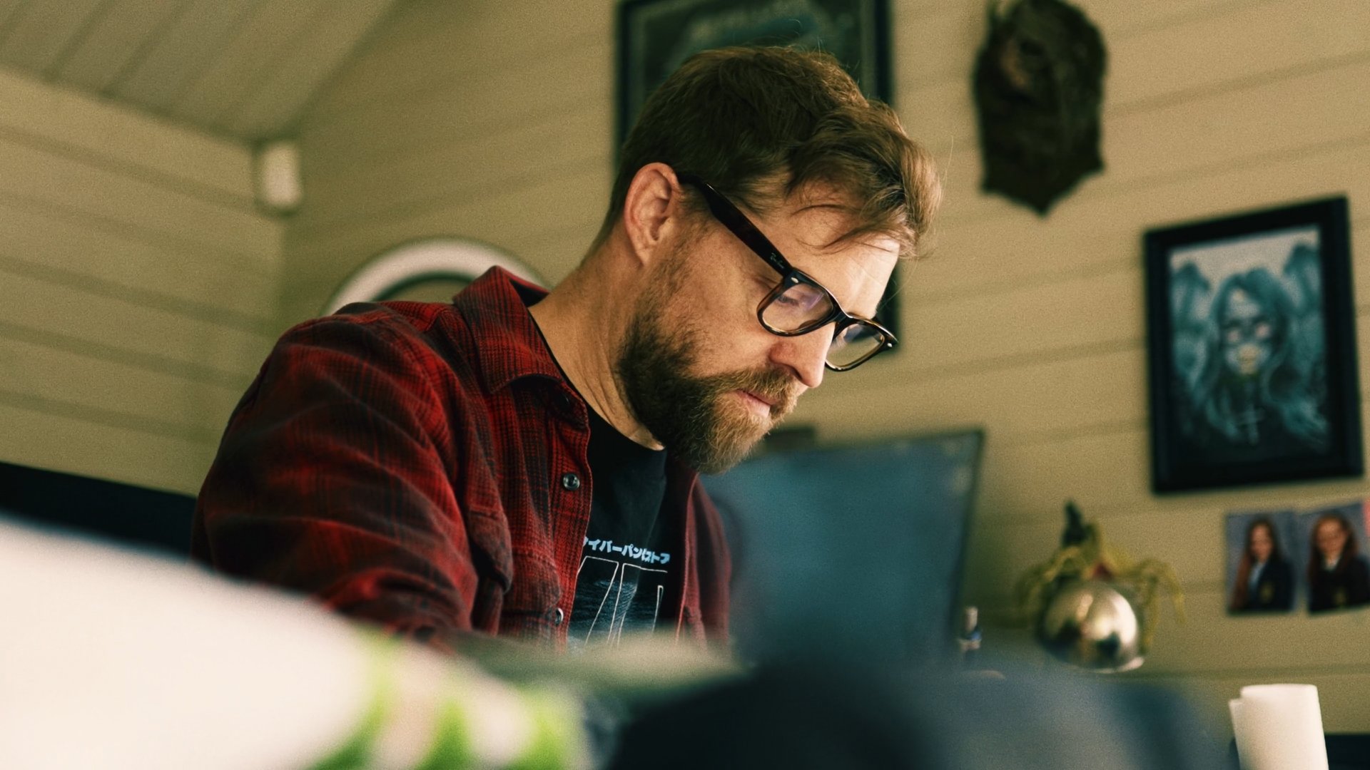 A man with glasses, a beard, and wearing a red plaid shirt is working or studying at a desk in a room decorated with framed pictures and paintings.
