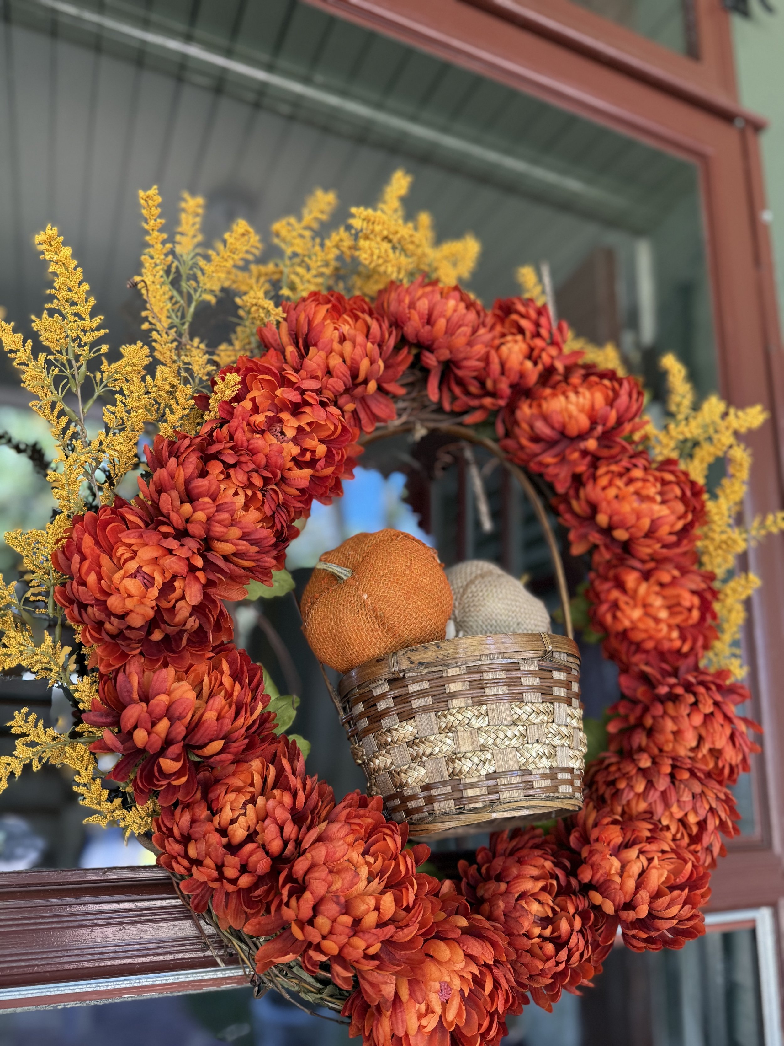 Fall wreath with orange and red flowers, yellow accents, and a small basket with fabric pumpkins hanging on a door.
