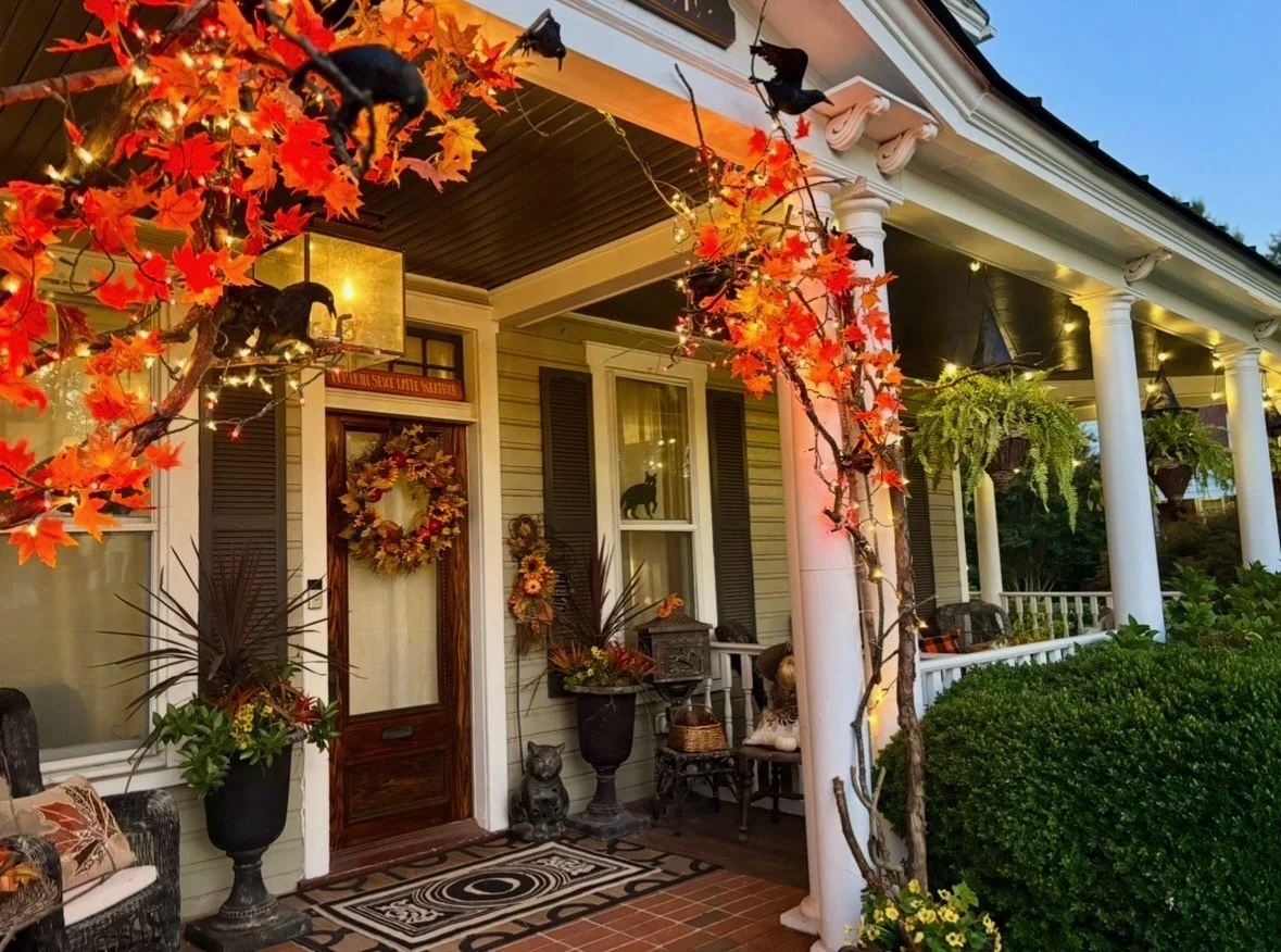 Decorated porch of a house with autumn-themed decorations, including a wreath, fall leaves, and string lights. Potted plants and a cat figurine are near the door, and a cozy seating area with chairs is visible.