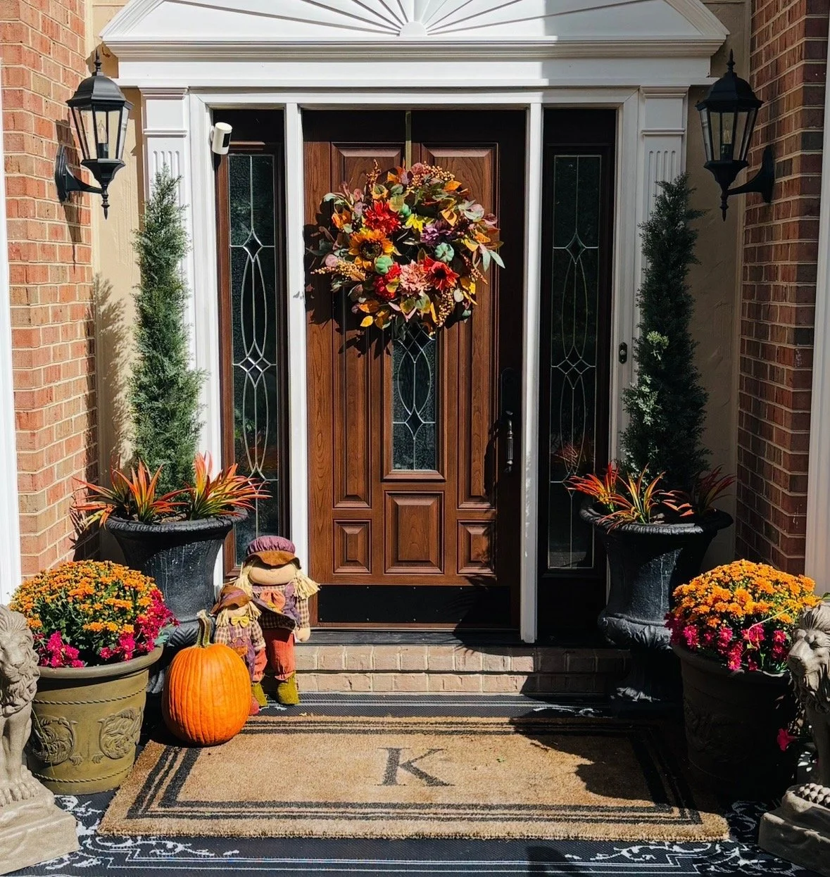 A front door decorated for fall with a floral wreath, potted plants, pumpkins, scarecrows, and a welcome mat.