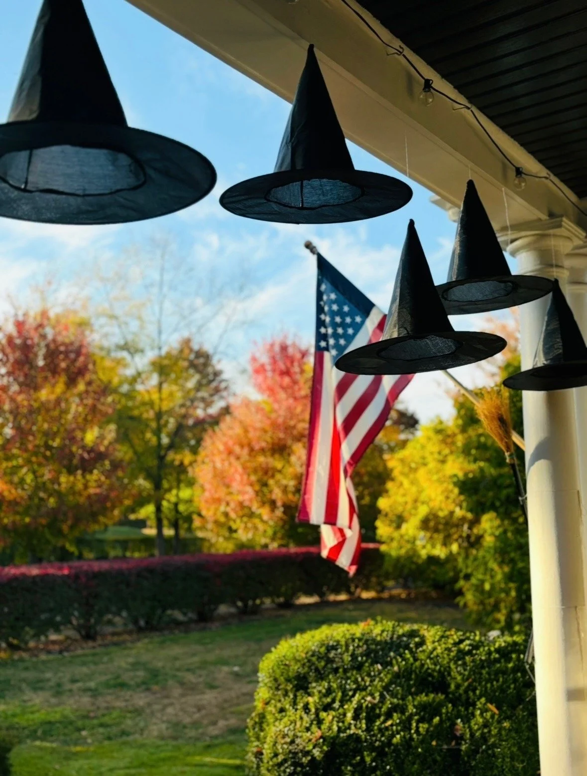 Halloween decorations hanging from a porch ceiling, featuring black witch hats and an American flag outside with trees showing fall foliage in the background.