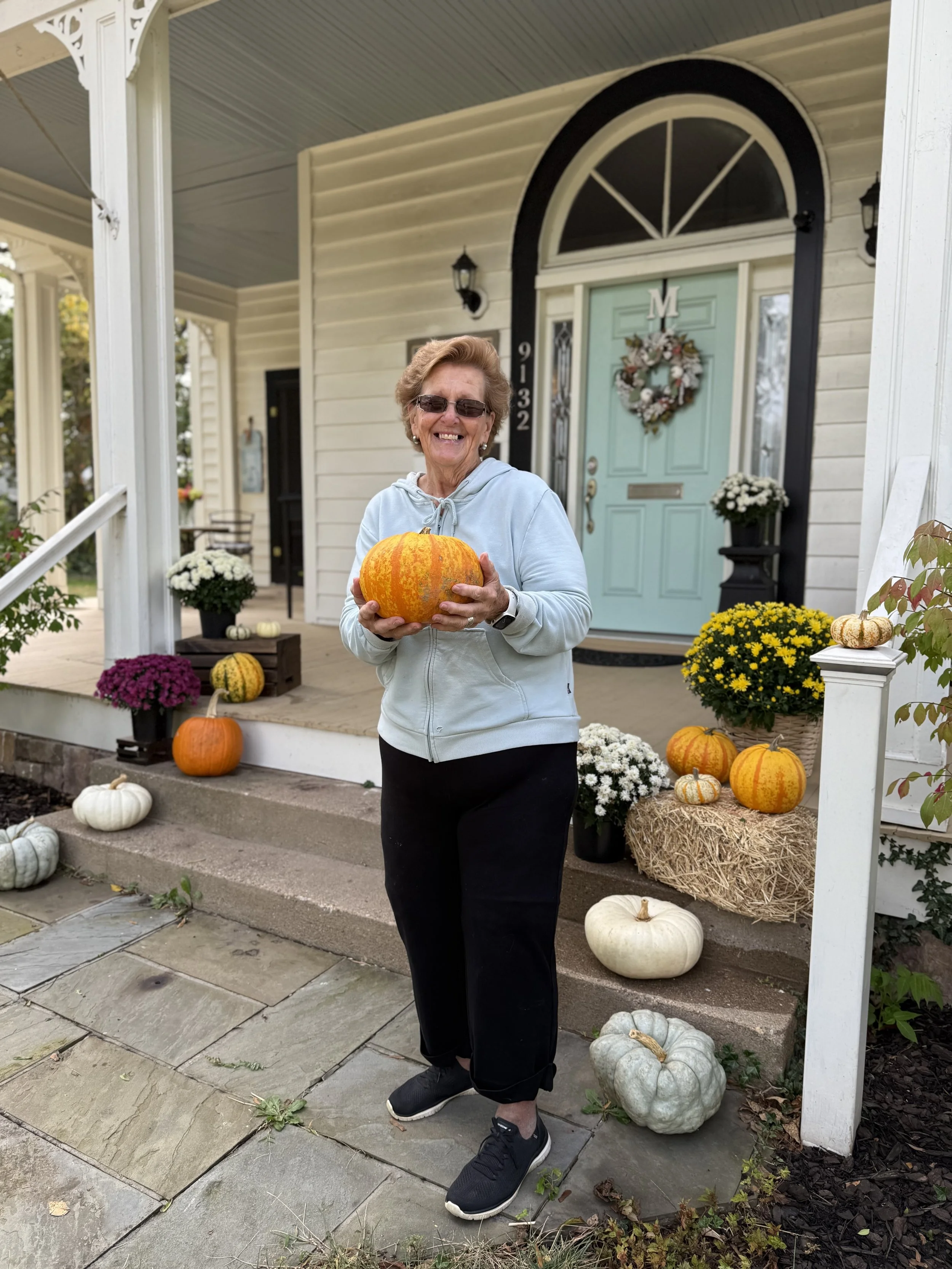 An elderly woman in a light gray hoodie and black pants holding a pumpkin, standing on the front steps of a house decorated with pumpkins and flowers for fall.