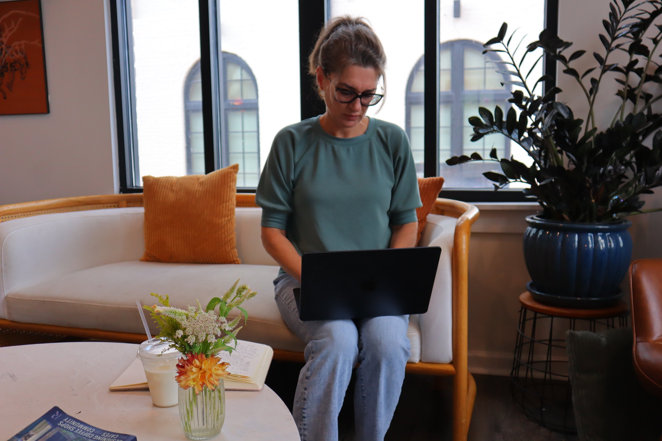 A woman with glasses and a gray-green shirt sitting on a white sofa with an orange pillow, looking at a laptop. There is a large window behind her with buildings outside. To her right, there is a large potted plant, and on the table in front of her, there is a vase with flowers and an open notebook.