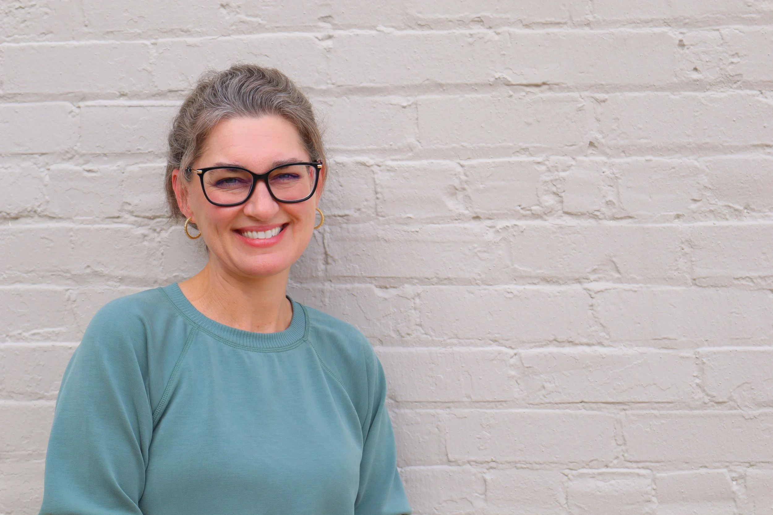A smiling woman with glasses and hoop earrings stands in front of a white brick wall.