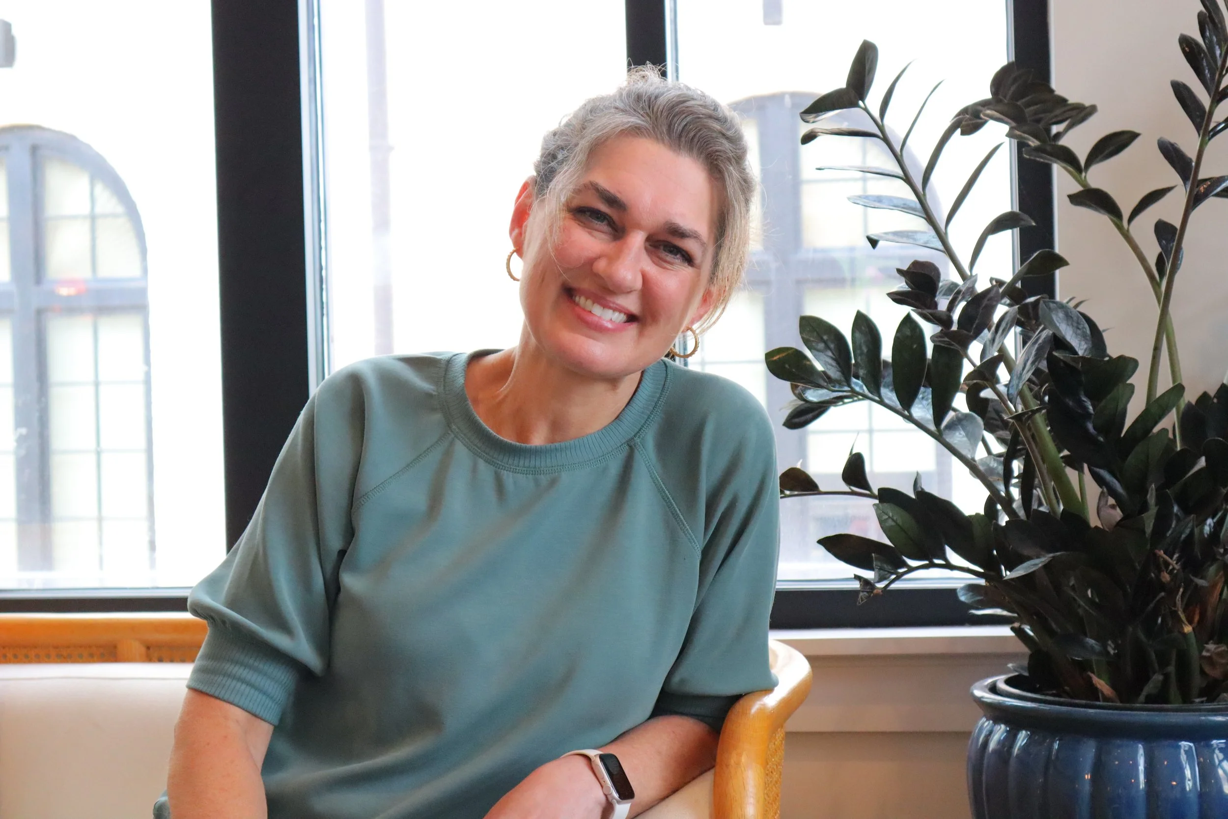 A smiling woman with gray hair tied back, wearing a teal top, seated indoors near a large window with sunlight. There is a large dark green-leafed plant in a blue pot beside her.