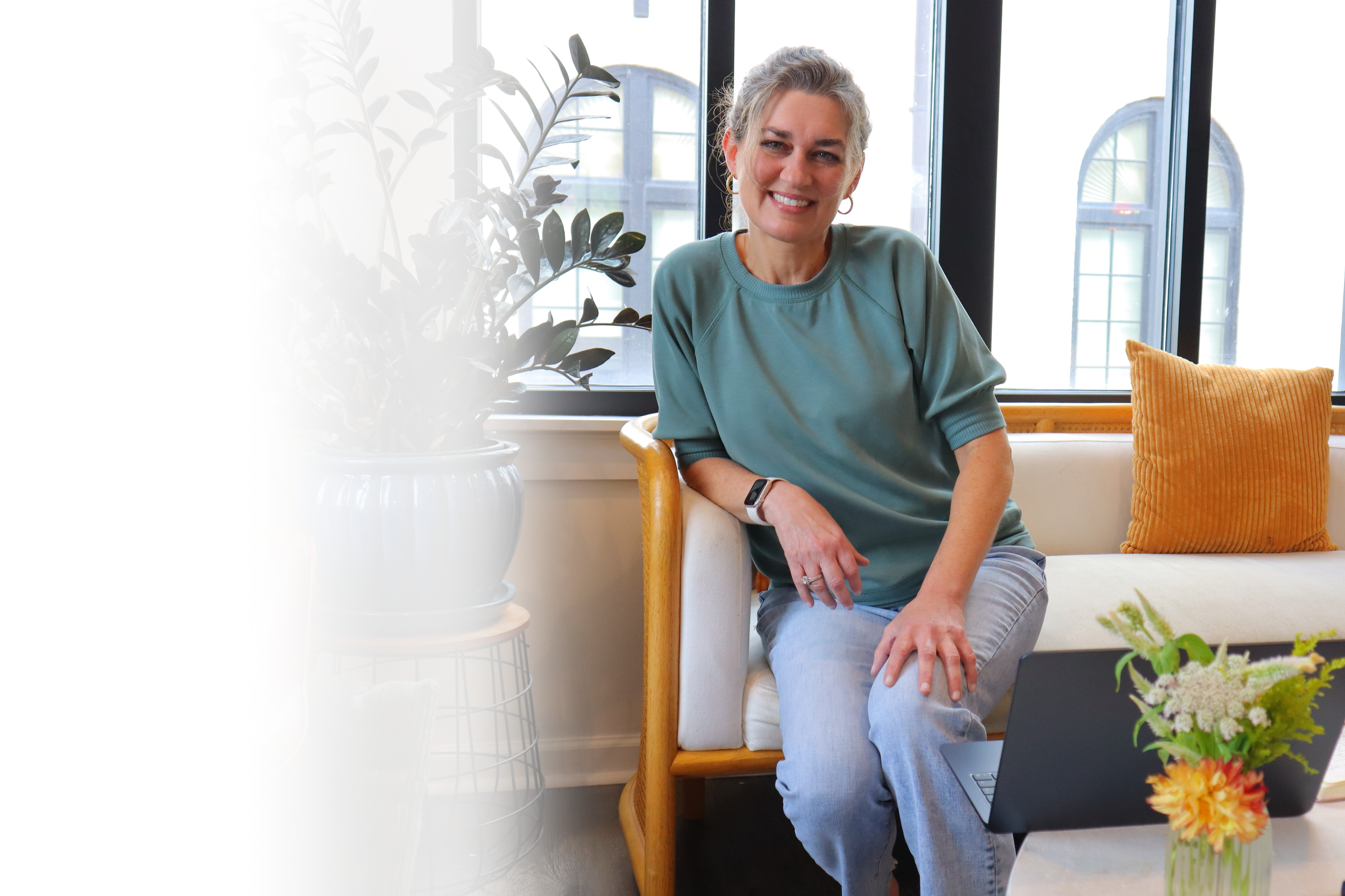 Smiling woman sitting on a wooden chair near a window with a large potted plant, a yellow cushion, and flowers on a table.