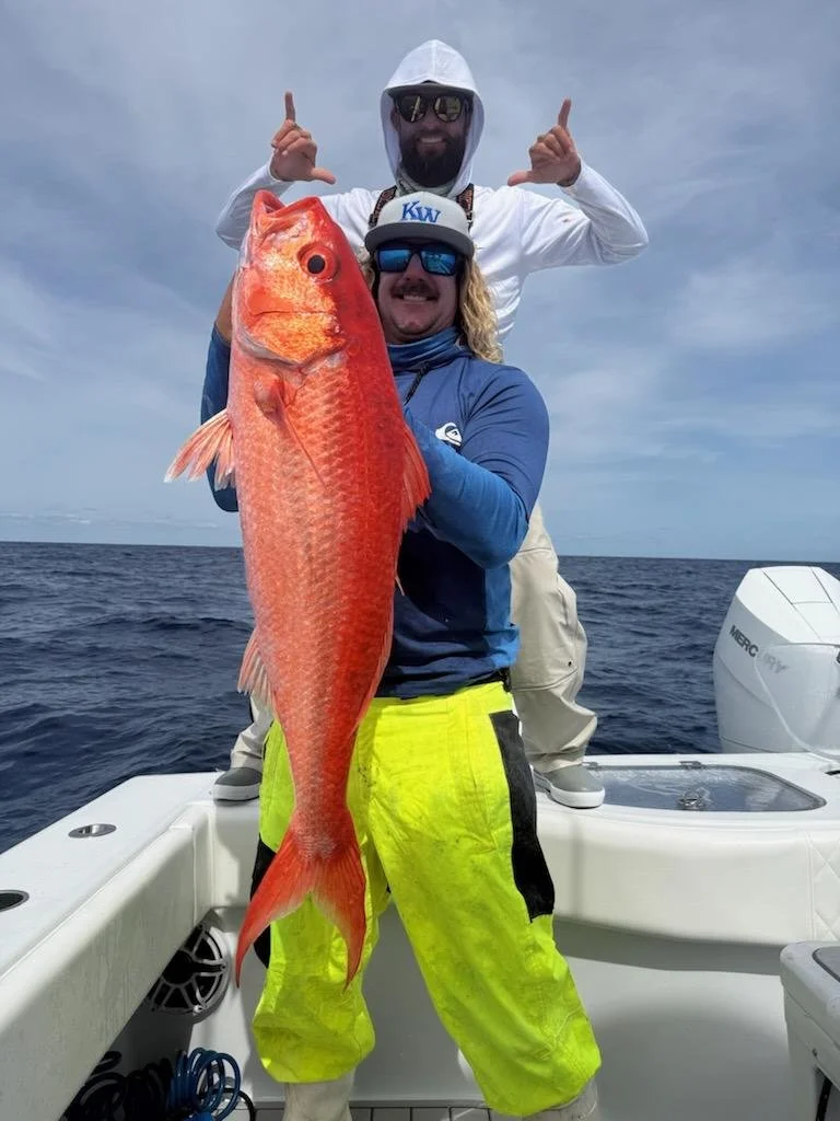 Two men on a boat holding a large red fish, with a blue sky and ocean in the background.