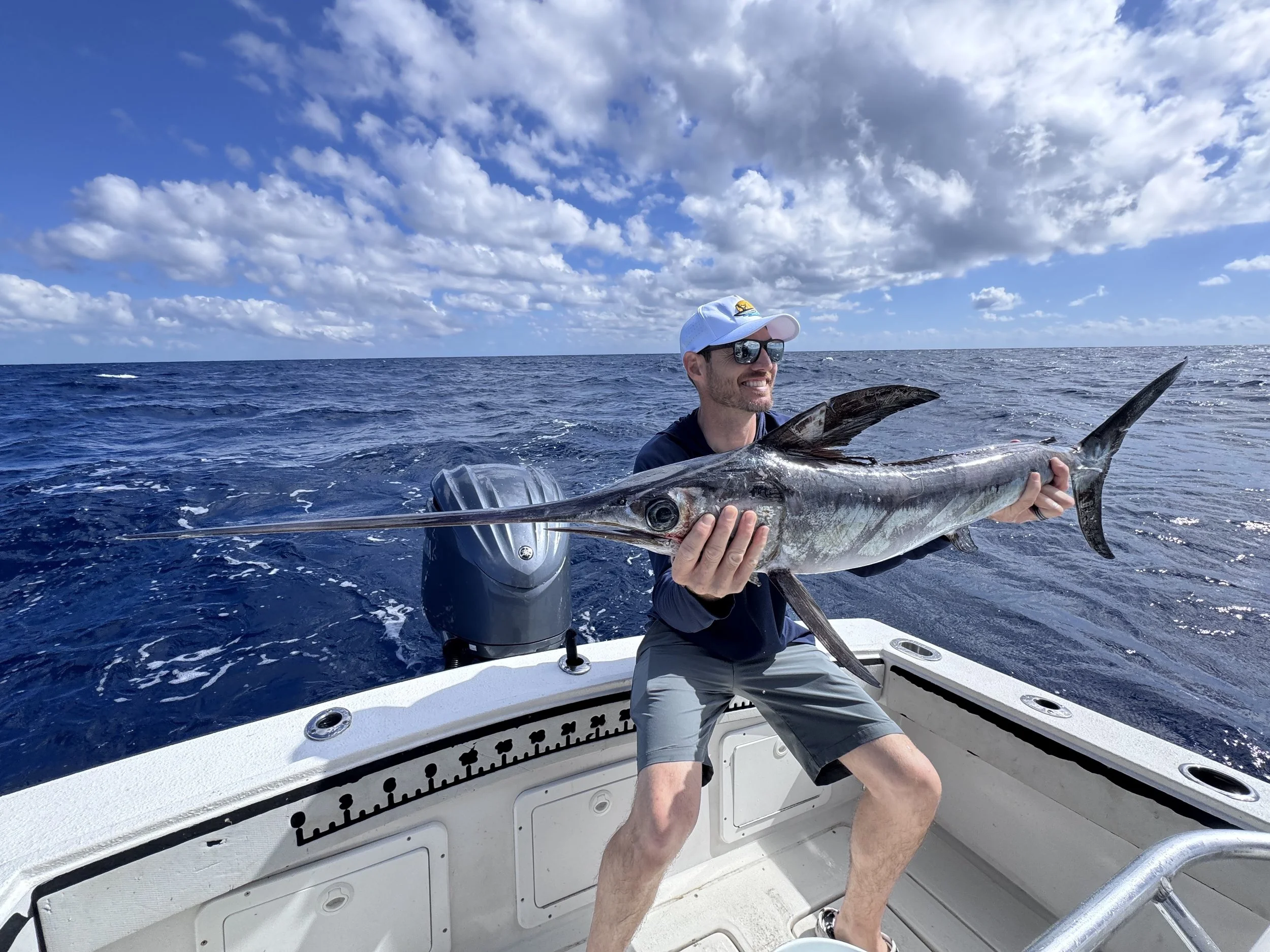 A man wearing sunglasses, a cap, and casual clothes, sitting on a boat and holding a large marlin fish with a long pointed bill, while smiling at the camera on a sunny day with blue sky and clouds over the ocean.