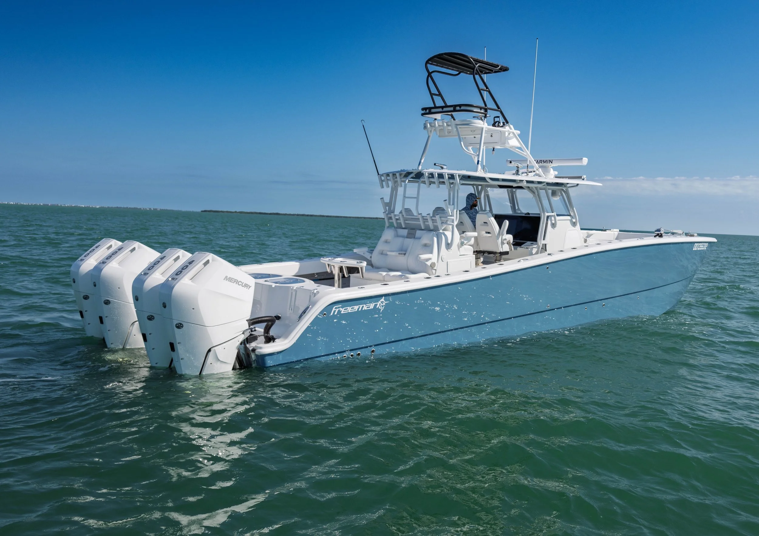 A white and blue Freeman boat with three Mercury outboard motors, floating on the ocean against a blue sky.