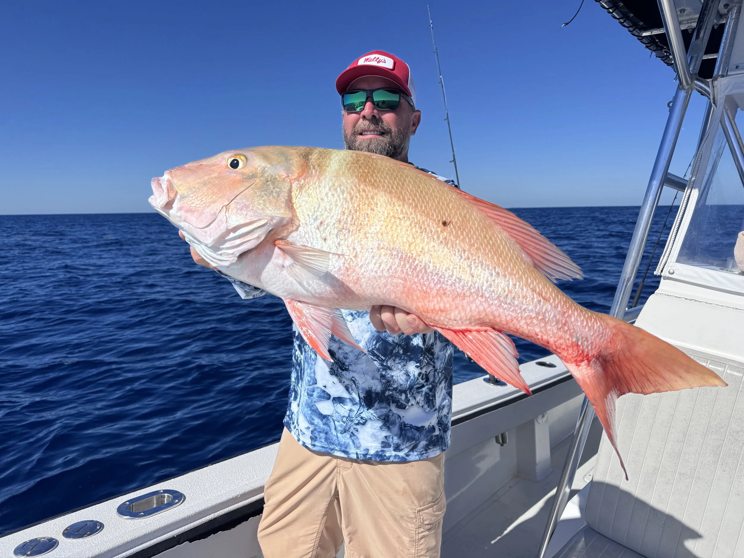 A man wearing sunglasses and a red cap holding a large pinkish-orange fish on a boat in the open ocean.
