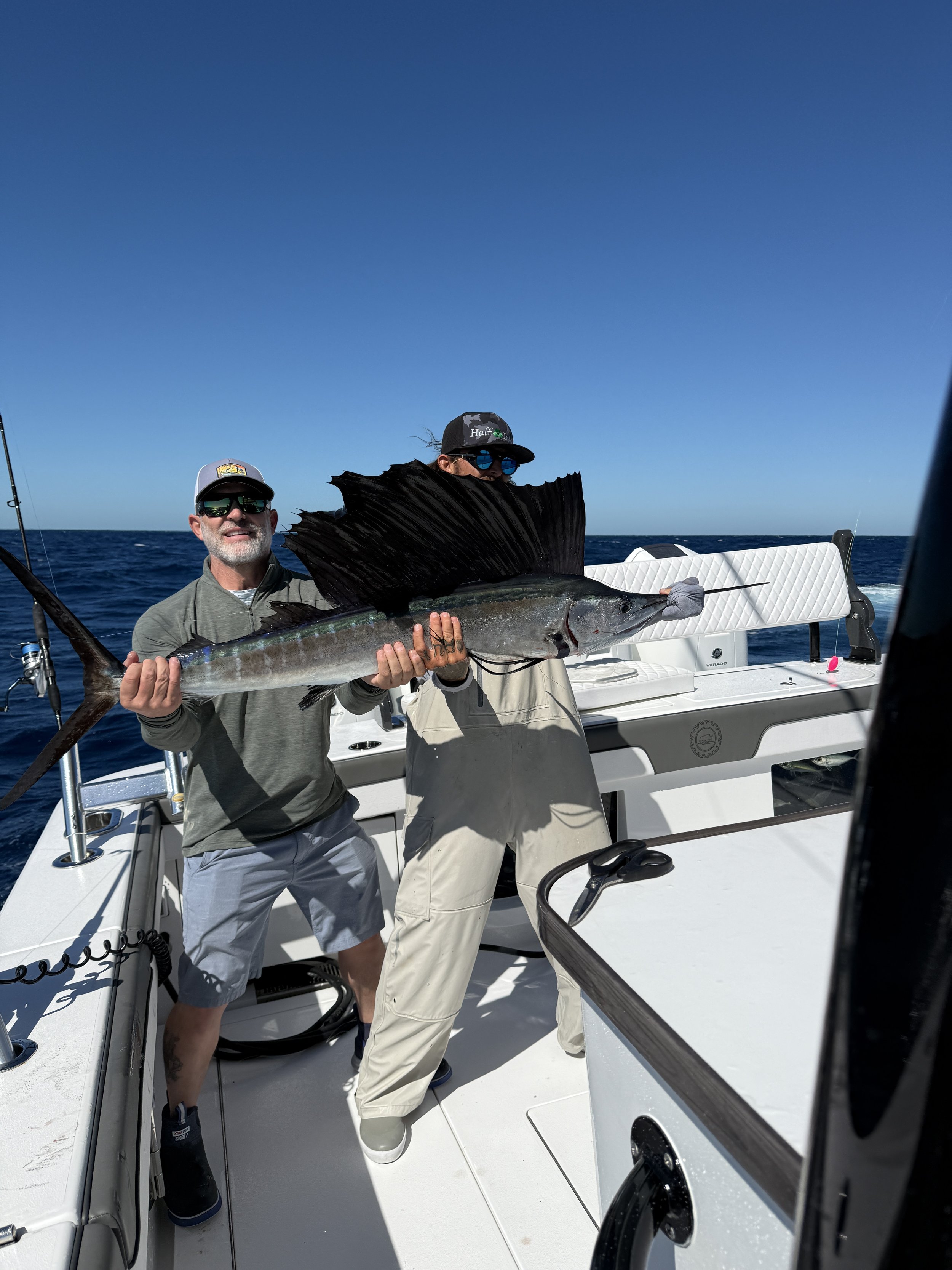 Two men on a boat holding a large fish with fins and a long body, against a clear blue sky and open ocean.