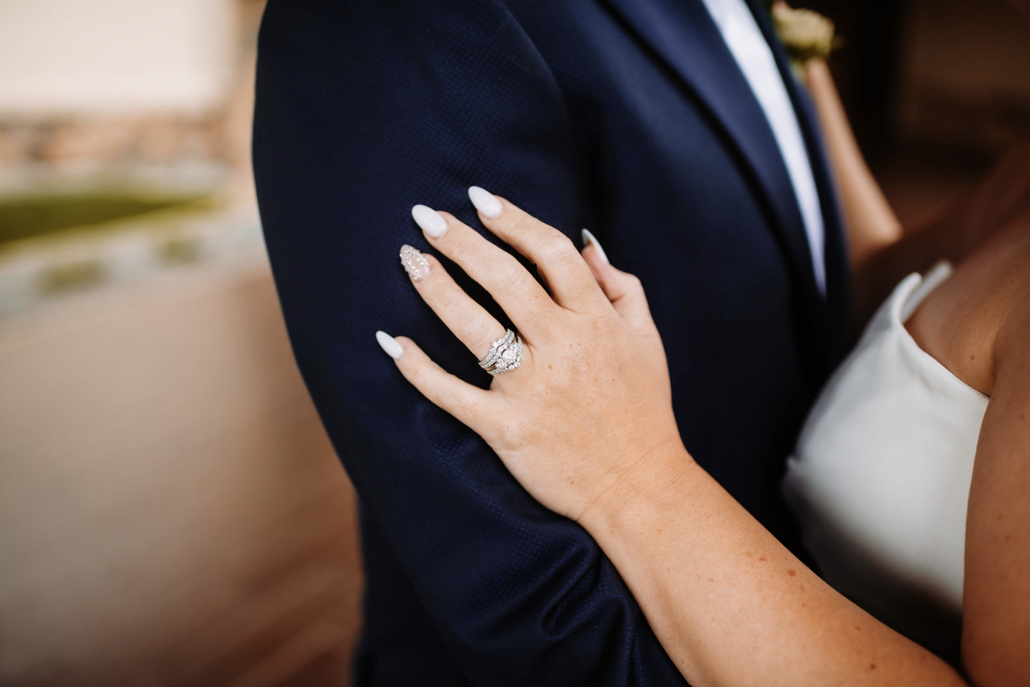 A woman with manicured nails and an engagement ring on her finger rests her hand on the shoulder of a man wearing a dark suit at a wedding or special event.