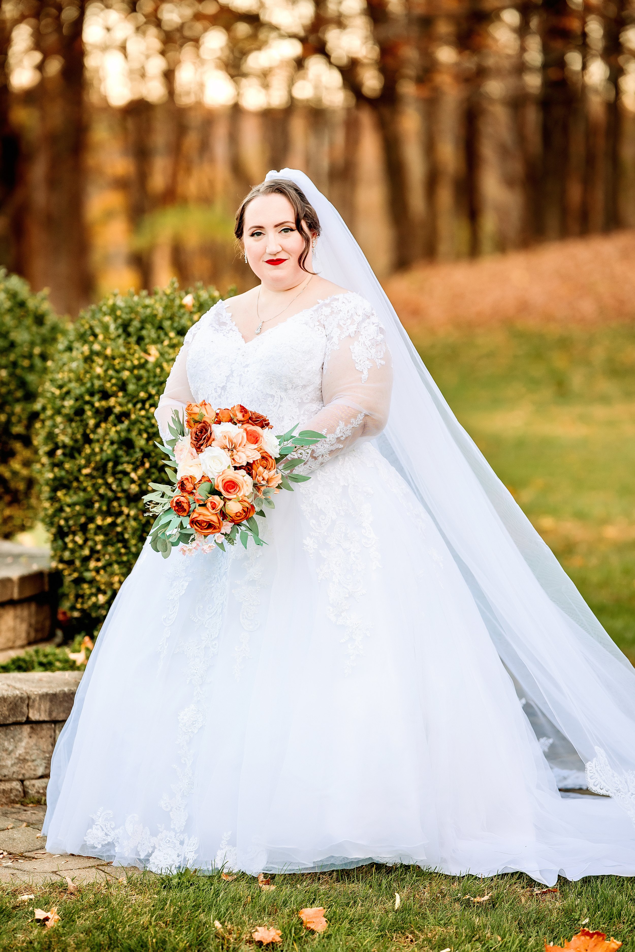 A bride in a white wedding dress holding a bouquet of orange, peach, and white flowers outdoors during autumn.