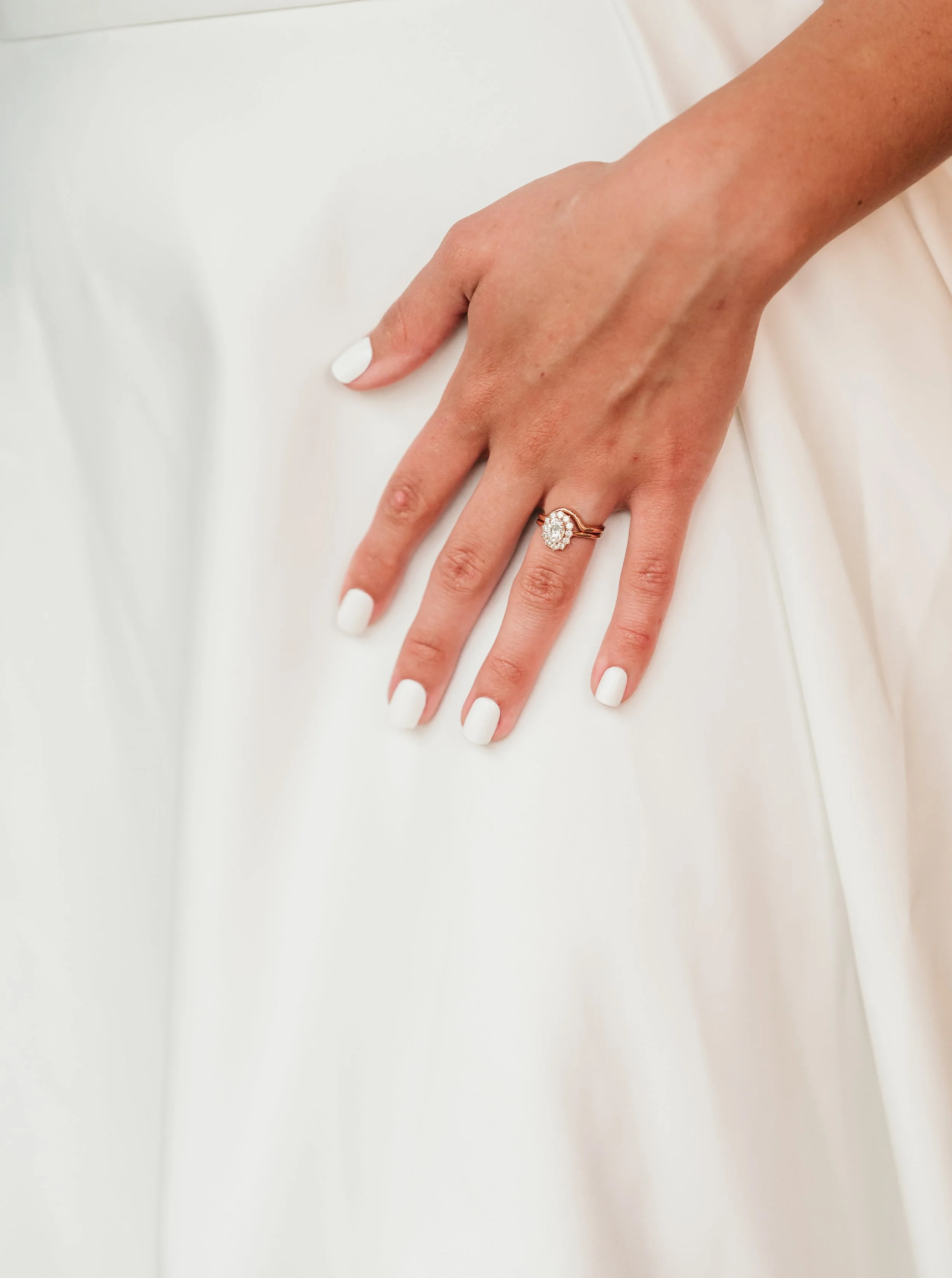 Close-up of a woman's hand with a diamond engagement ring, resting on a white fabric surface.