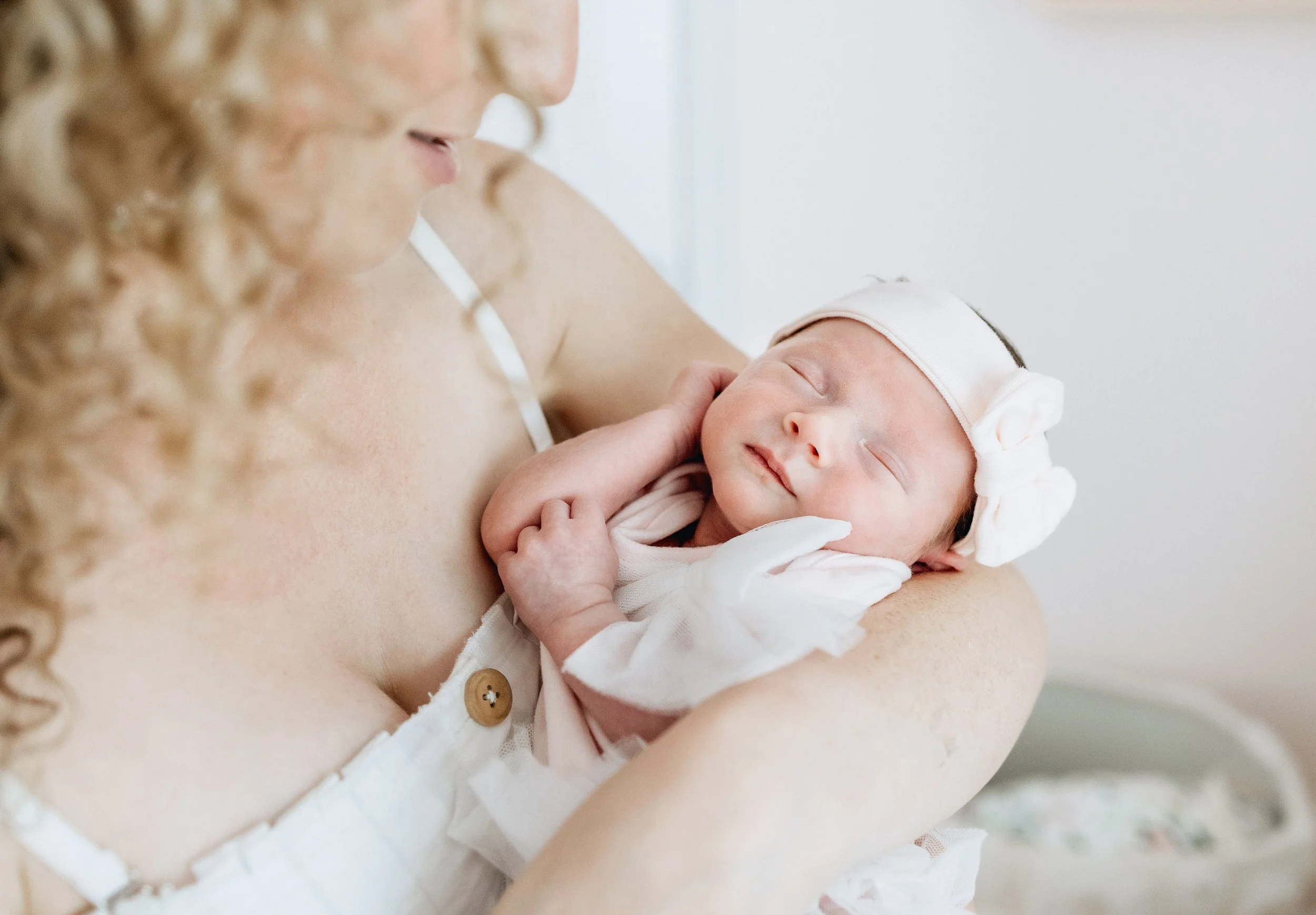 A woman holding a sleeping baby against her chest, with her face partially visible and curly hair, in a soft-lit setting.