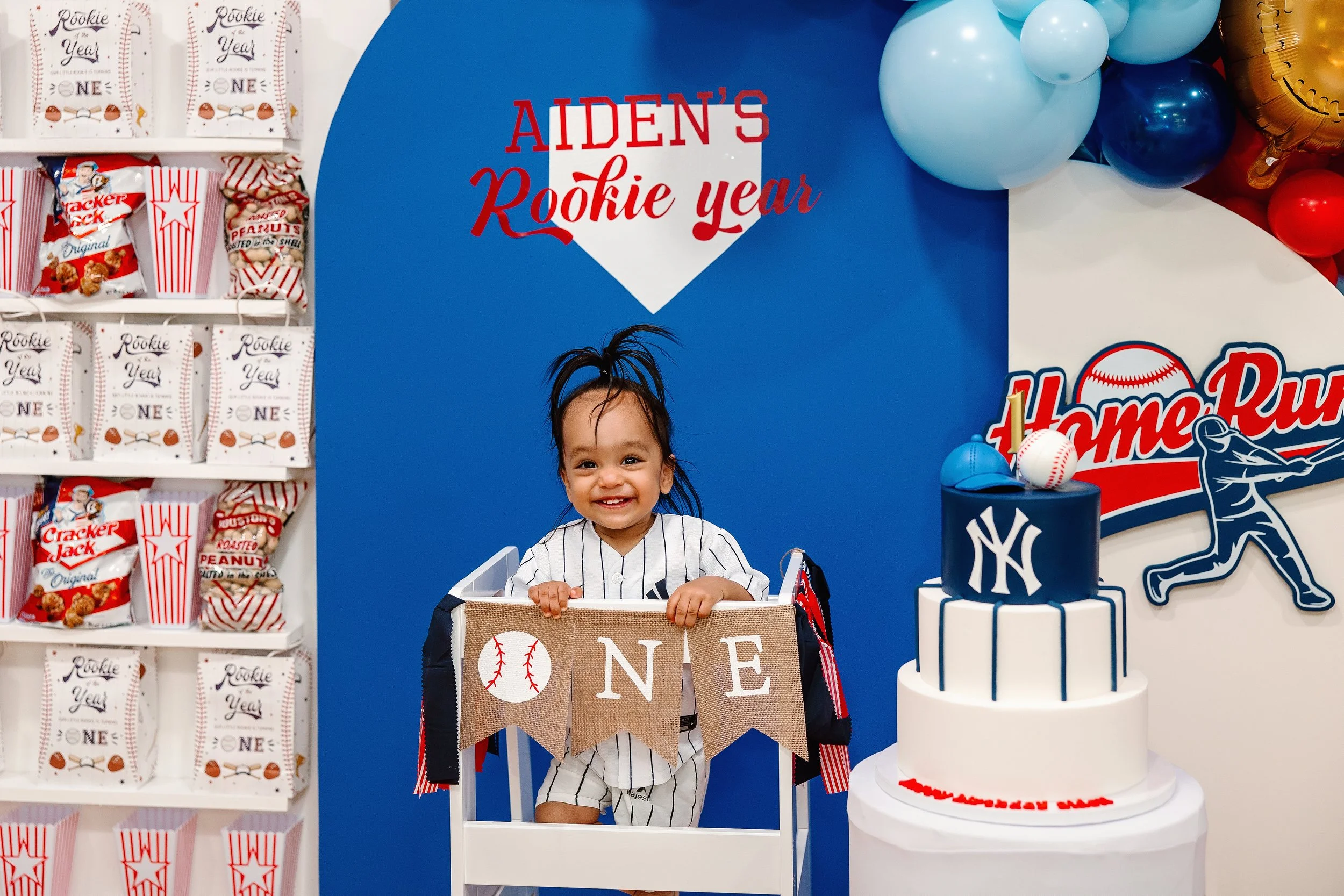 A young child celebrating their first birthday with a baseball-themed party, wearing a baseball jersey, surrounded by baseball decorations, a birthday cake with baseball and New York Yankees logos, and a backdrop that reads 'Aiden's Rookie Year'.