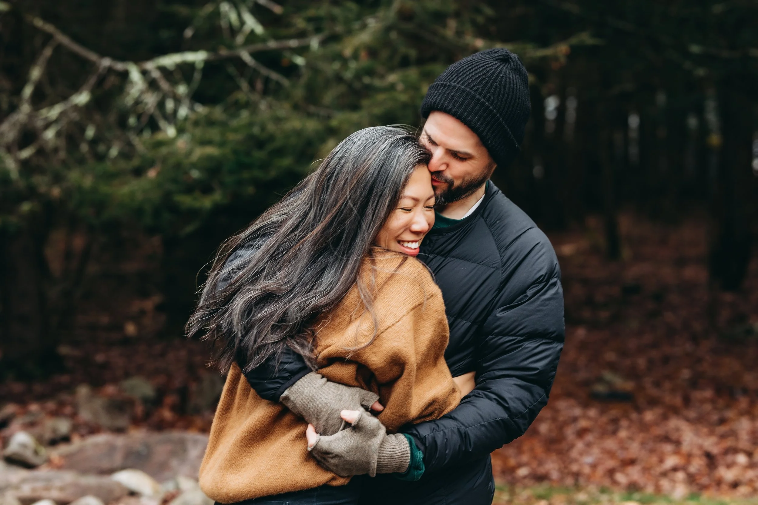 A couple hugging outdoors in a forest, smiling and showing affection.
