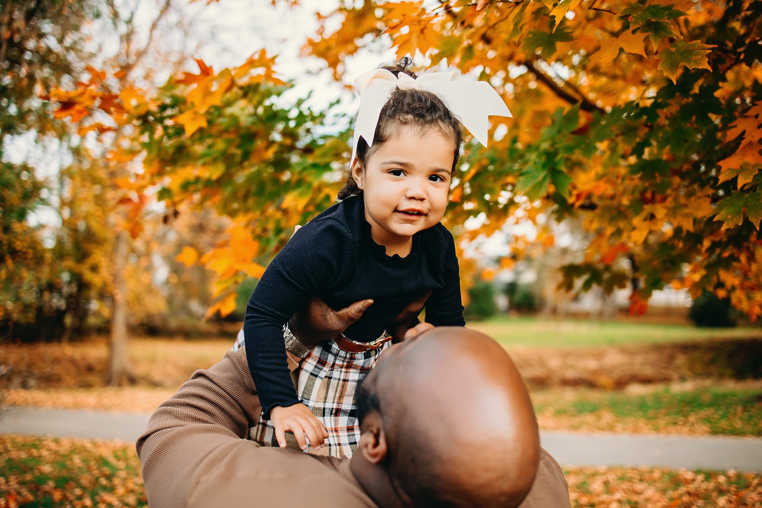 A young girl with a white bow in her hair smiling as she sits on a man's shoulders during fall, surrounded by orange and yellow leaves.