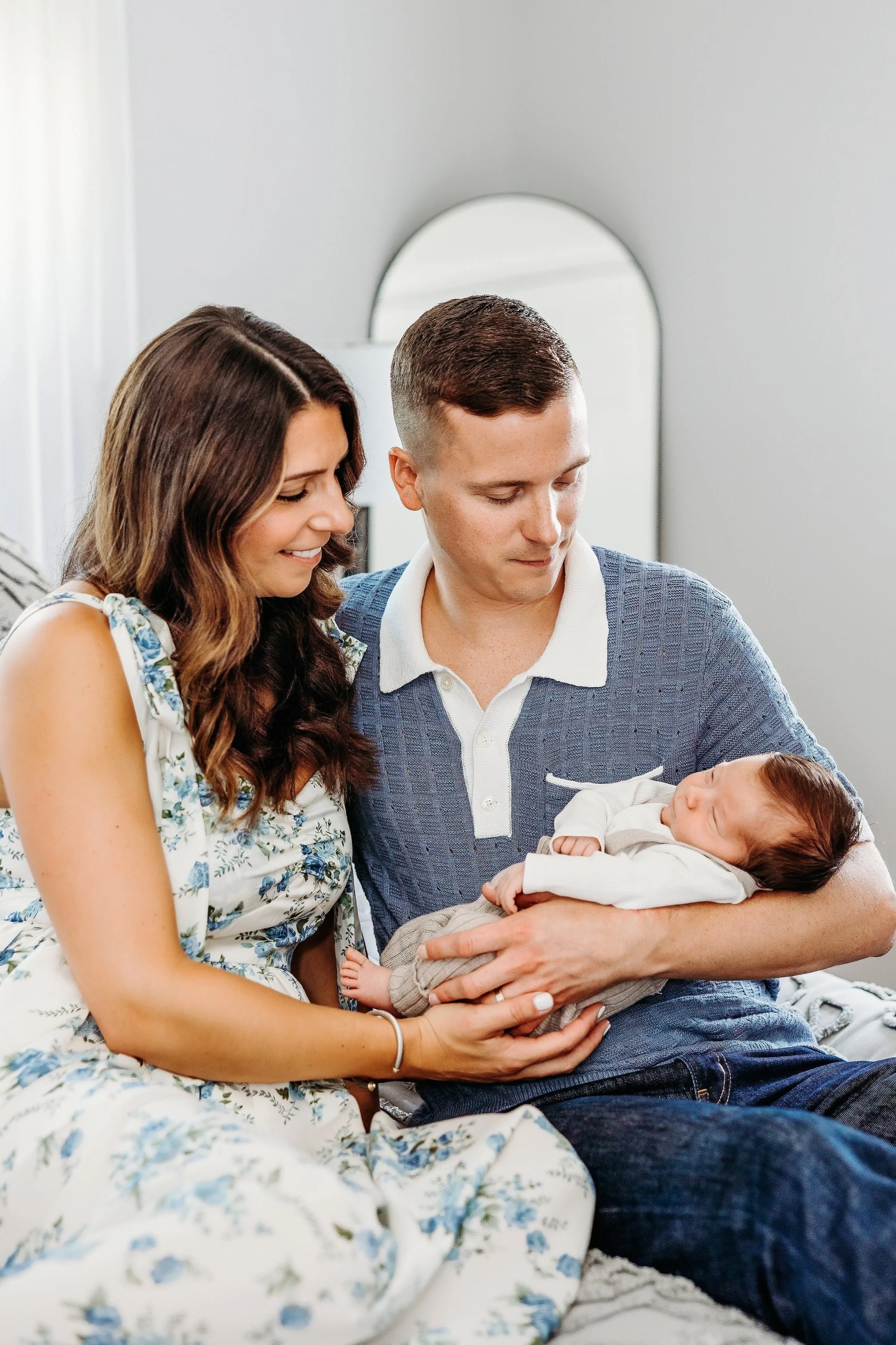 A young couple sitting on a bed, holding and looking at a newborn baby in a cozy bedroom with a mirror in the background.