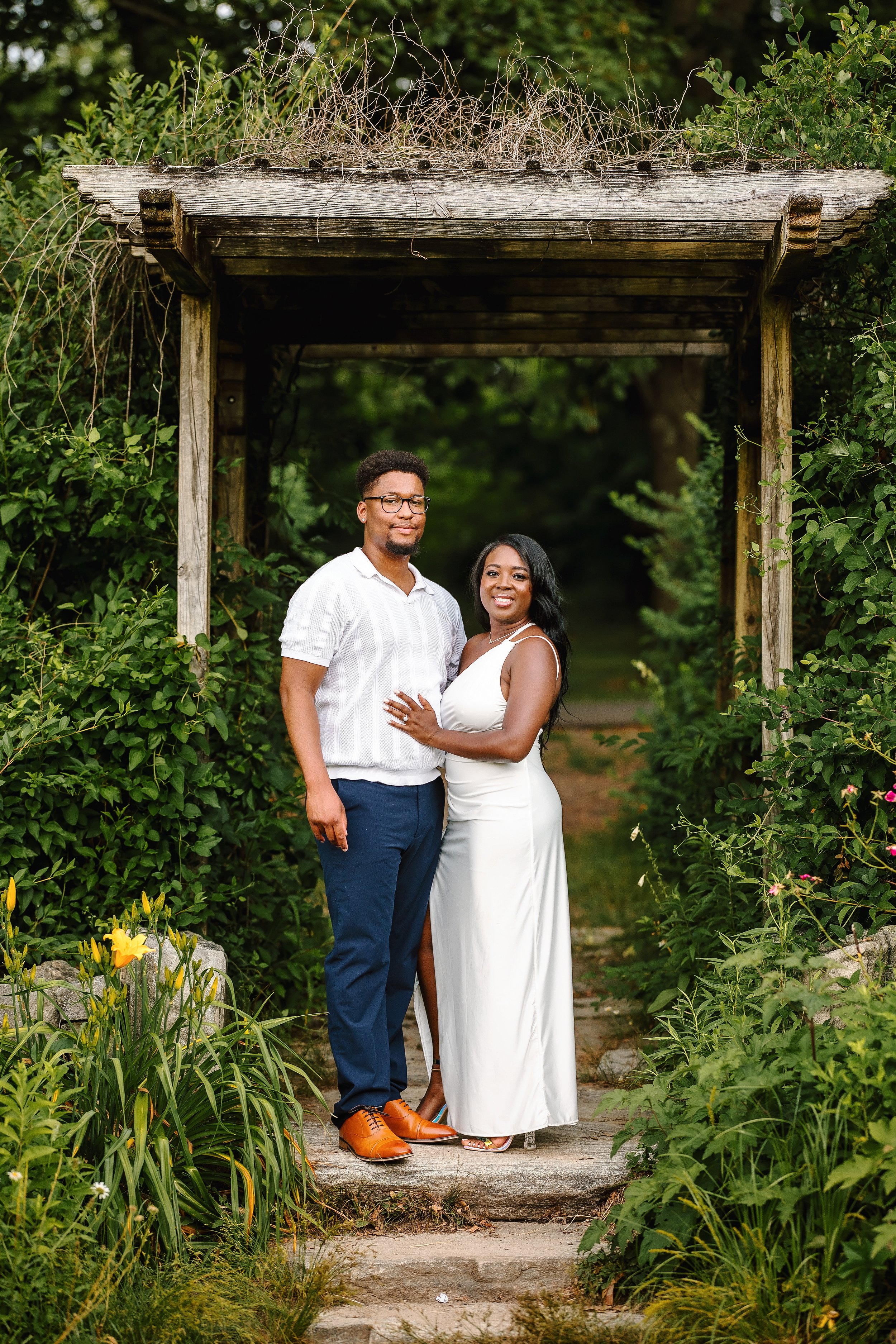A couple standing under a wooden archway surrounded by green foliage and yellow flowers, dressed in formal attire, smiling at the camera.
