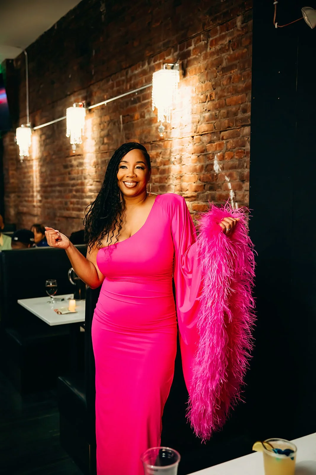A woman in a pink gown holding a matching pink feathered stole, smiling in a restaurant with brick walls and hanging chandelier lights.