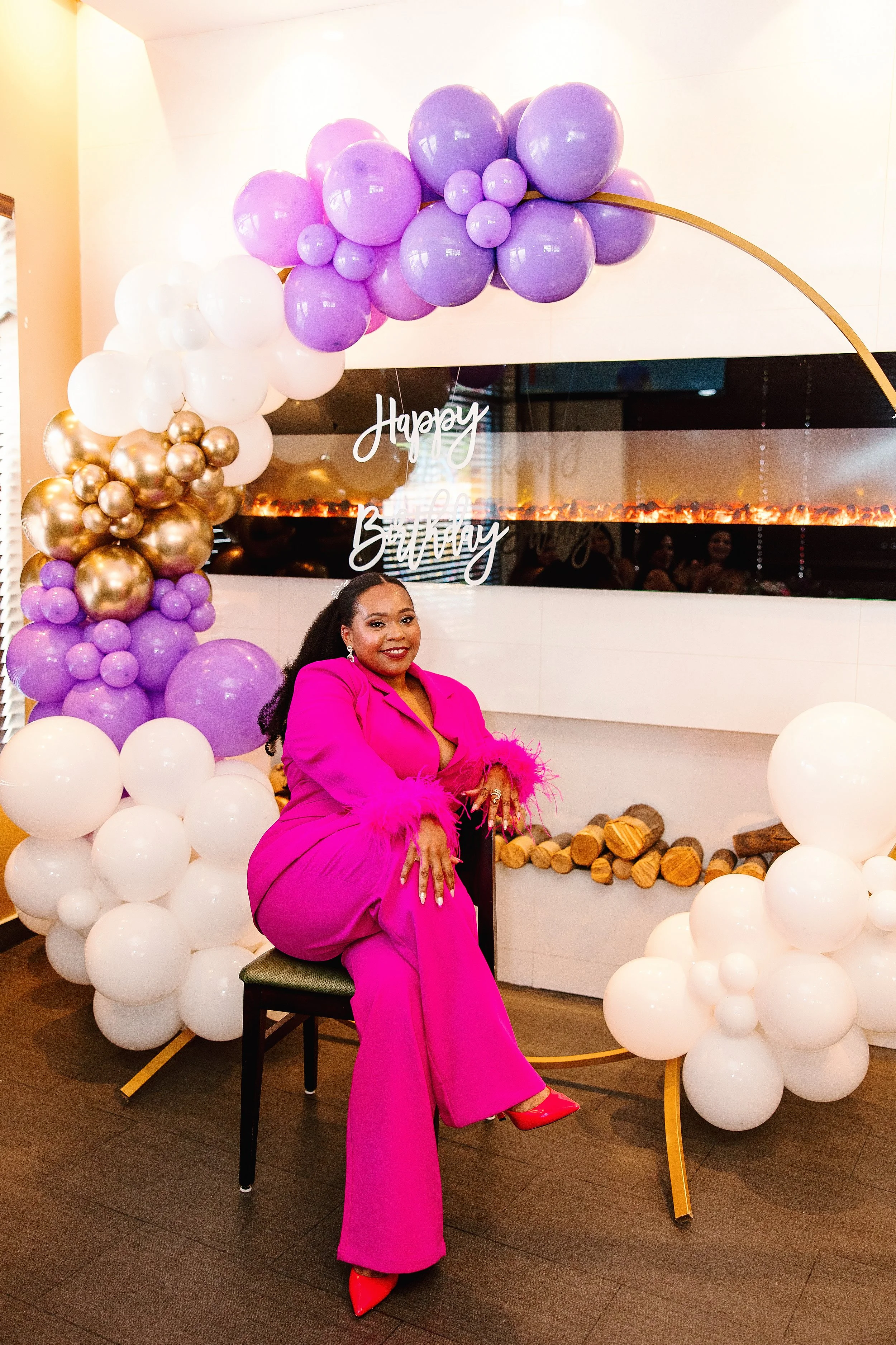 Woman in bright pink outfit sitting on a chair at a birthday celebration, surrounded by purple, white, and gold balloons, with a "Happy Birthday" sign in the background.