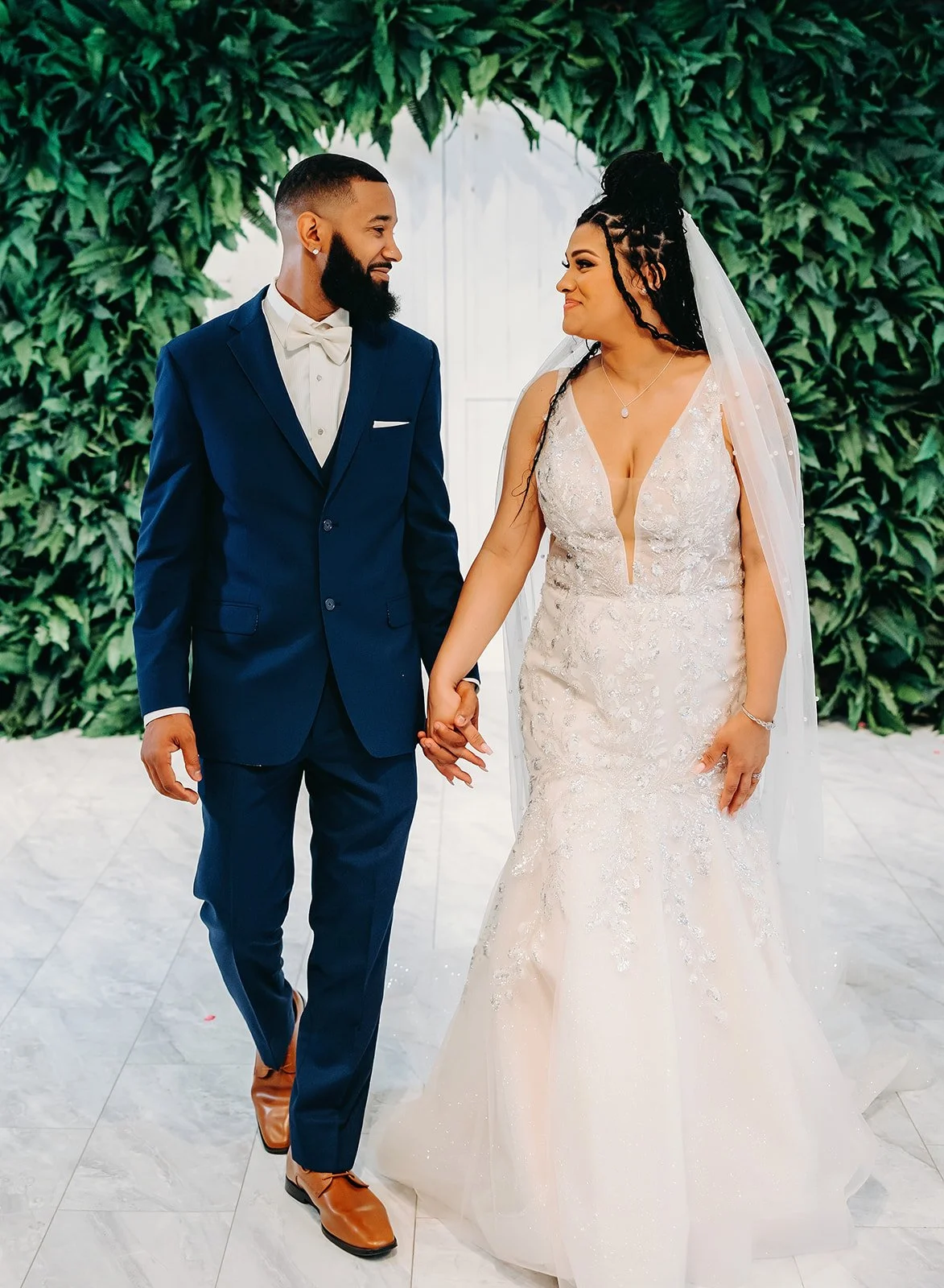 A bride and groom holding hands, looking at each other, standing indoors with a green leafy backdrop.