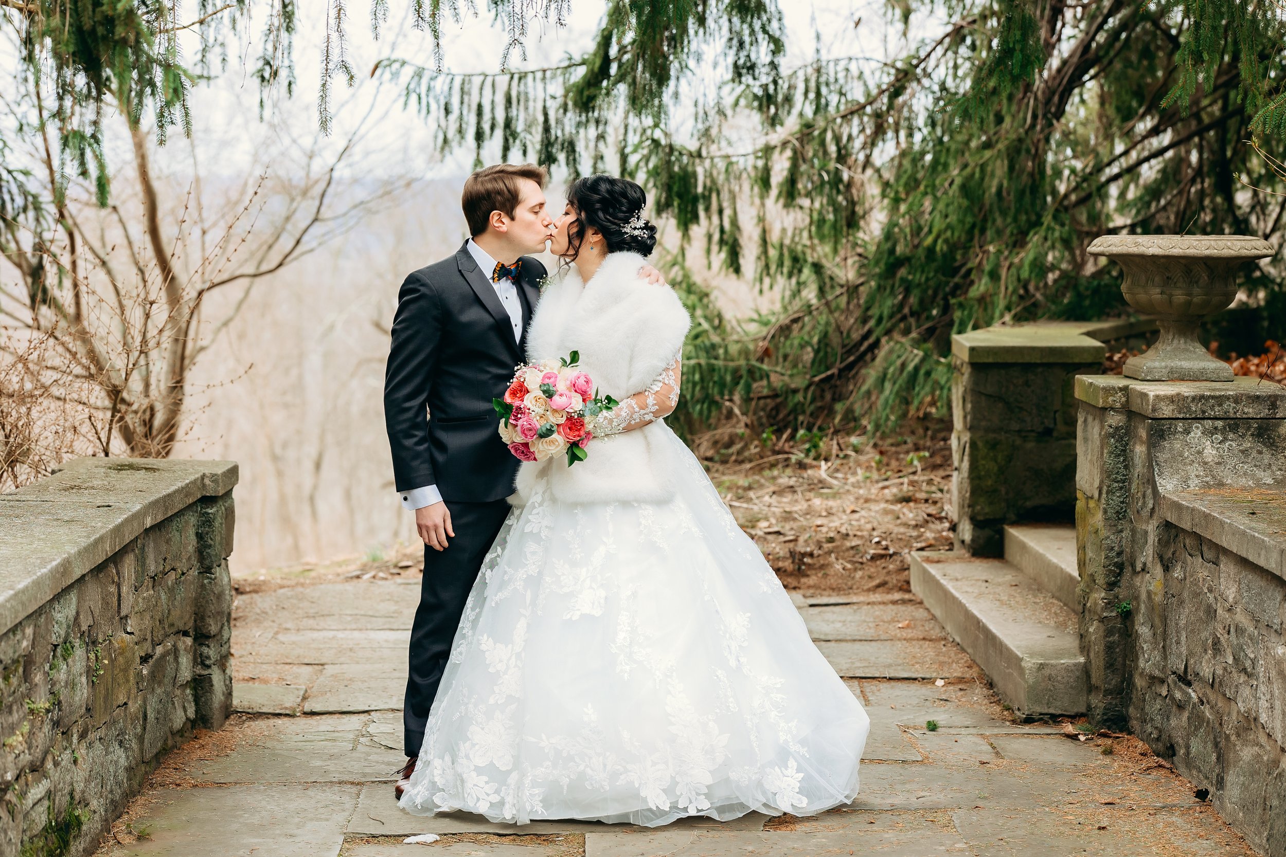 A newlywed couple sharing a kiss outdoors, the bride in a white wedding gown holding a bouquet of pink and white roses, and the groom in a black suit and bowtie, surrounded by trees and stone steps.