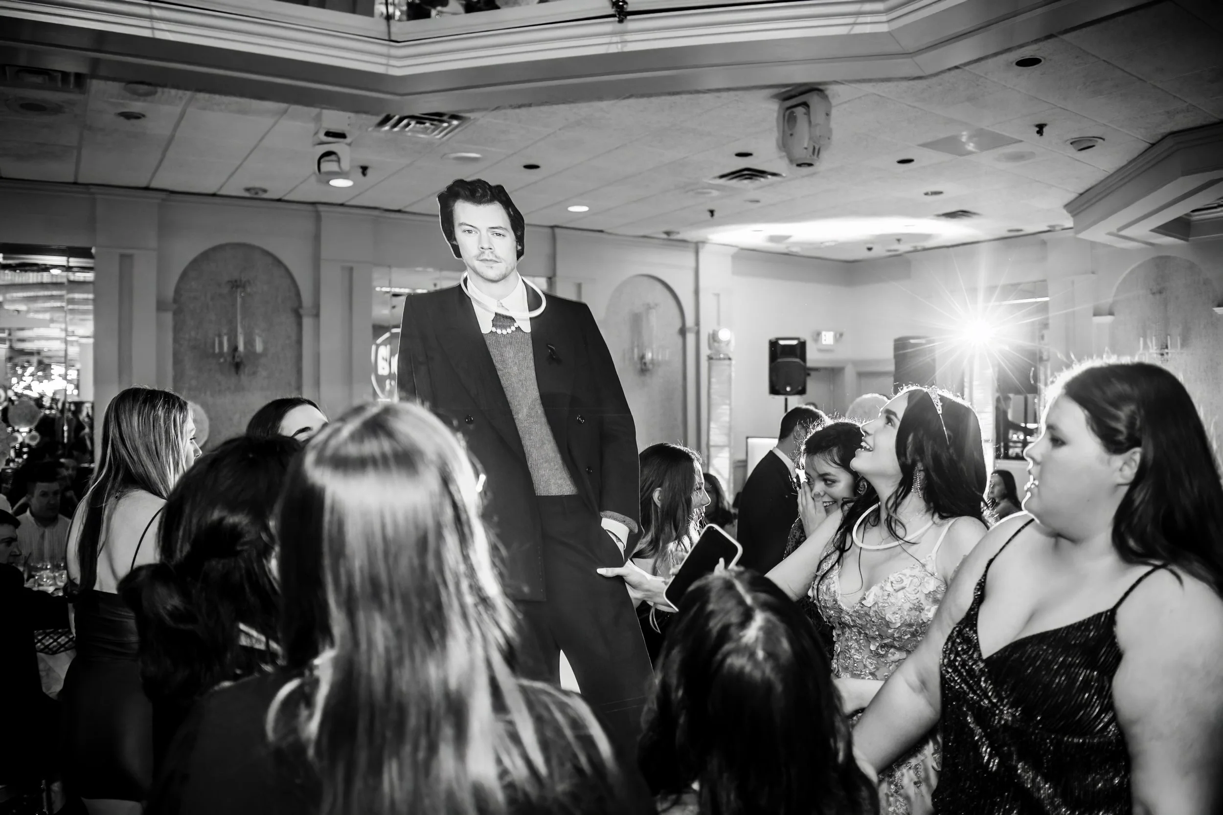 A black and white photograph of a lively social gathering with a crowd of women and a man, some women are smiling and engaging, in an indoor venue with ceiling lights and decorative walls.