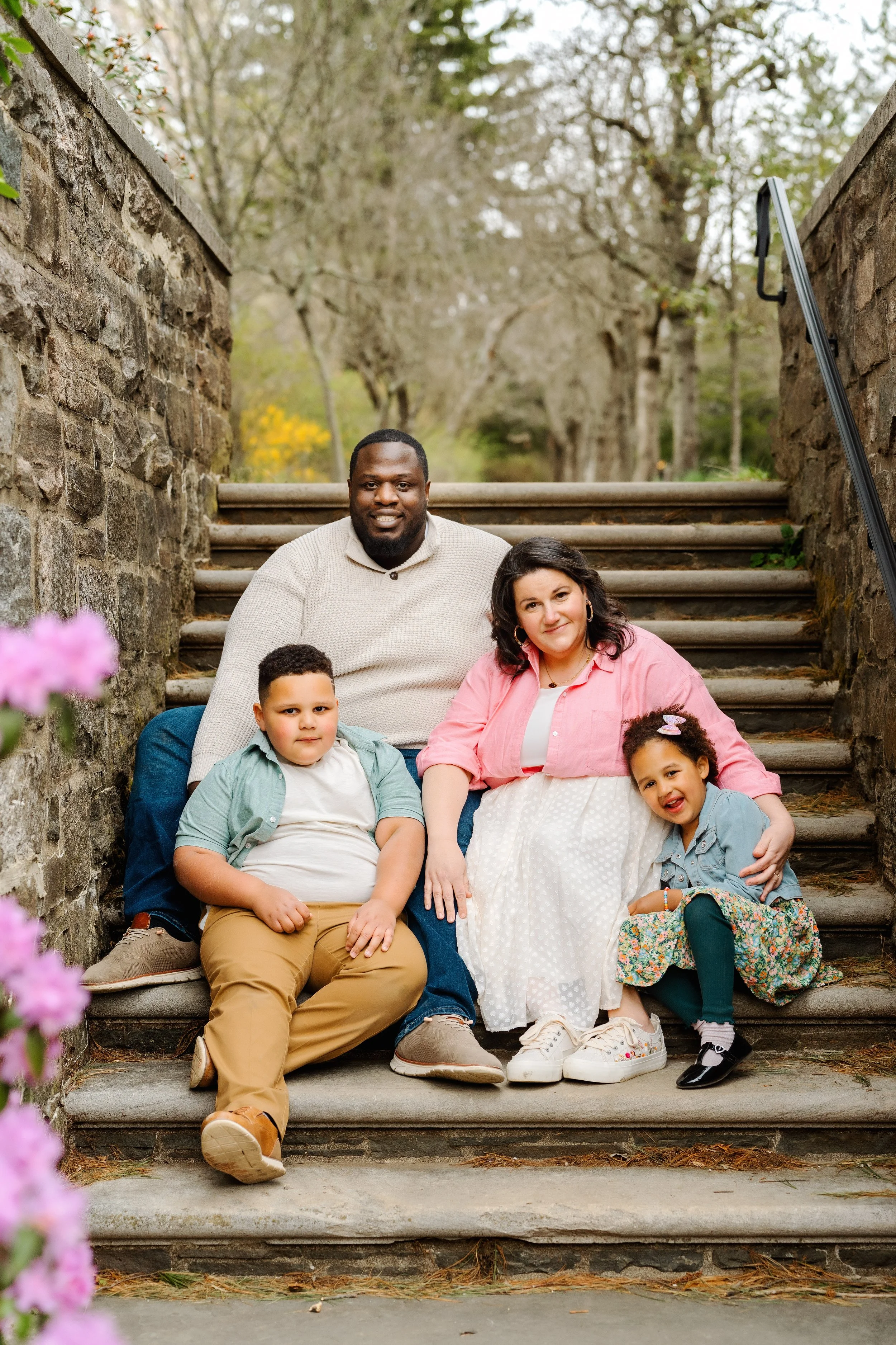 A diverse family of five sitting on outdoor stone stairs, surrounded by trees and pink flowers, smiling at the camera on a spring day.