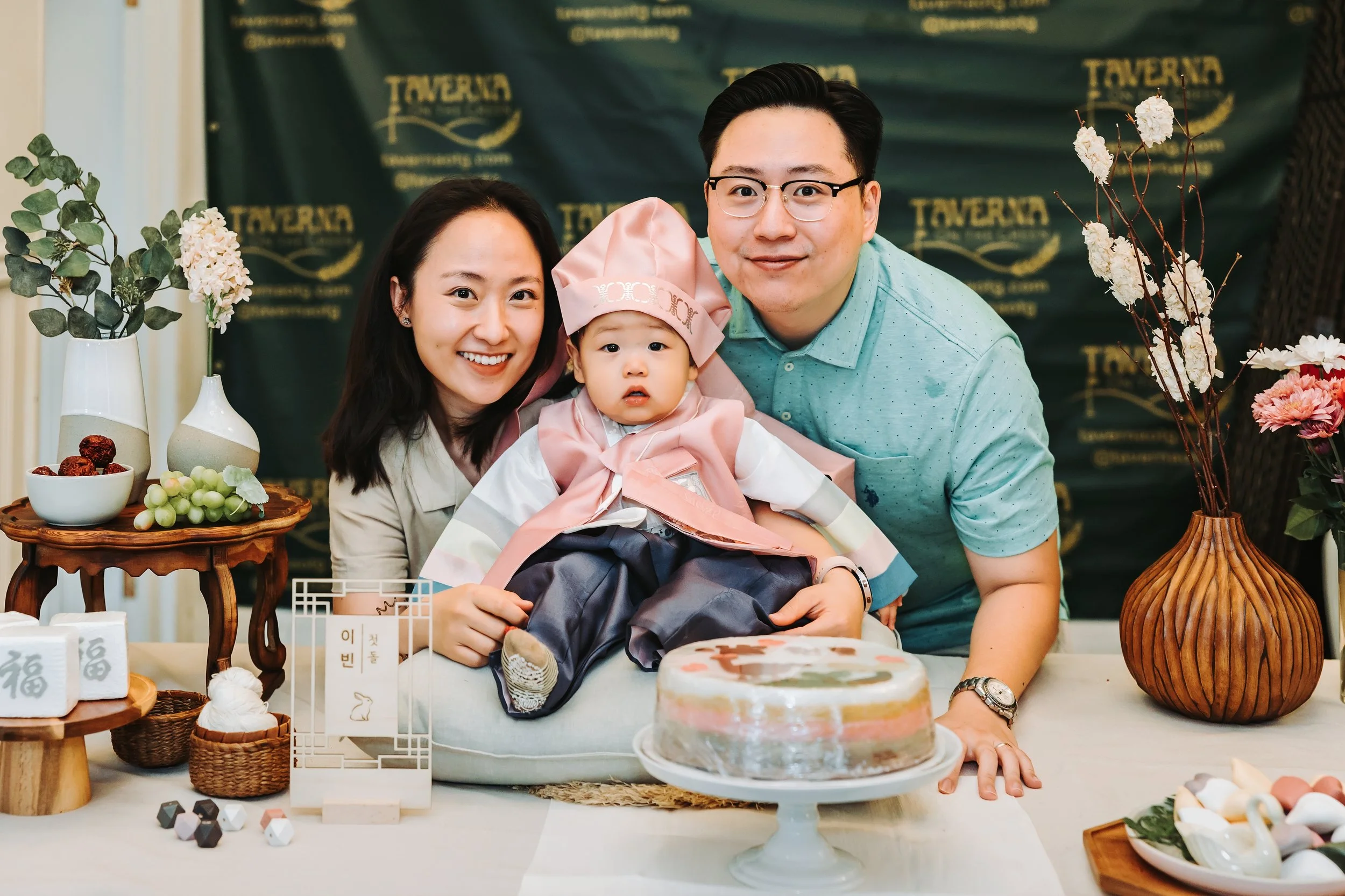 Family celebrating child's first birthday with cake, surrounded by traditional decorations and flowers.