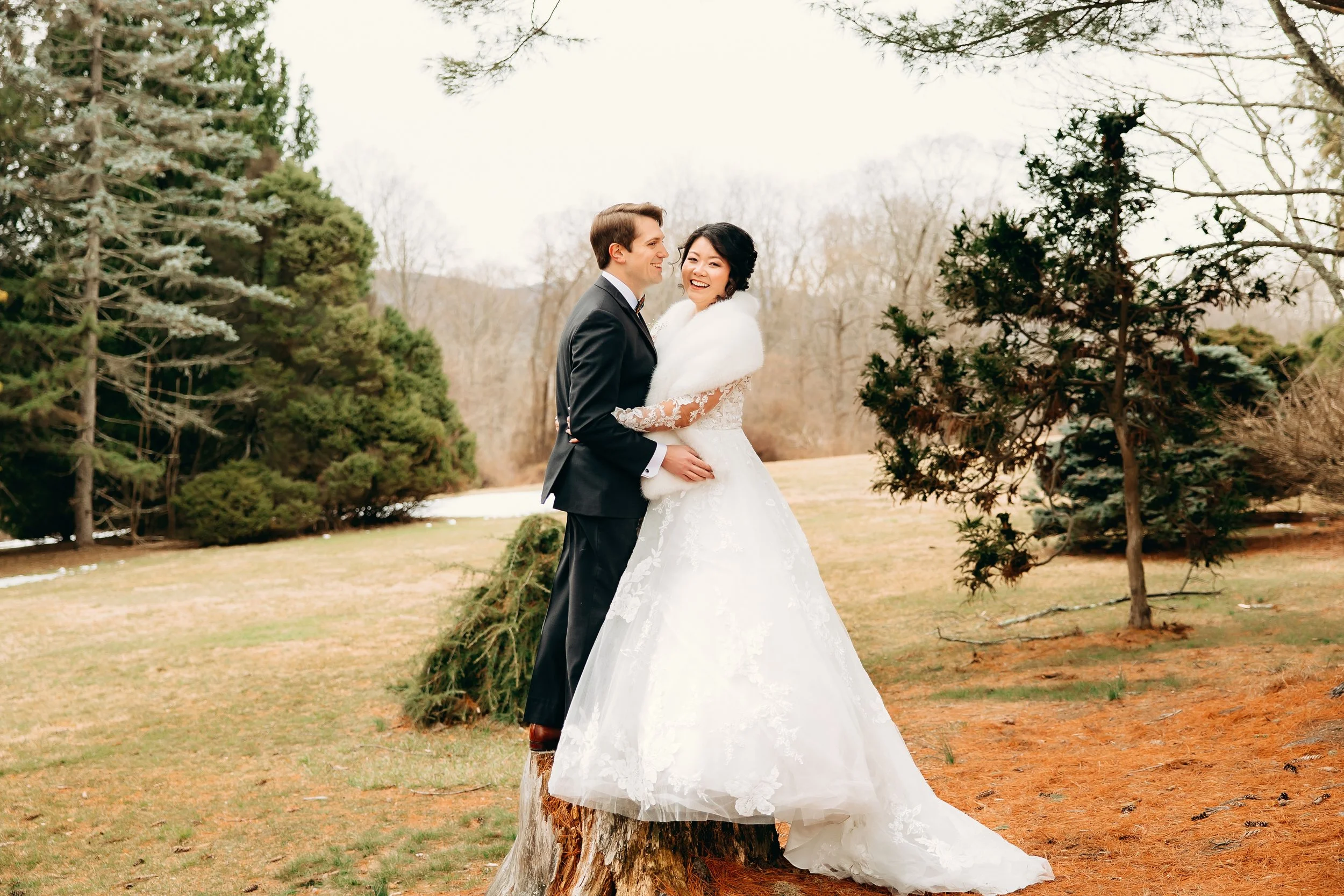 A newlywed couple in wedding attire standing outdoors on a tree stump, embracing and smiling, with trees and a partly cloudy sky in the background.