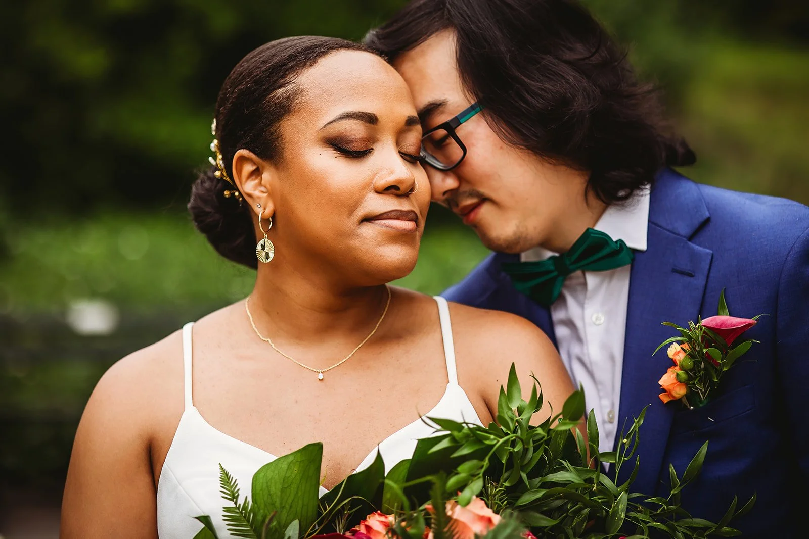 A couple facing each other with foreheads touching, outdoors during daytime, with the woman holding a bouquet of flowers. The woman has dark hair in a bun with decorative hairpins, wearing earrings and a necklace, dressed in a white sleeveless top. The man has long dark hair, glasses, wearing a blue suit with a white shirt, green bow tie, and a boutonniere.