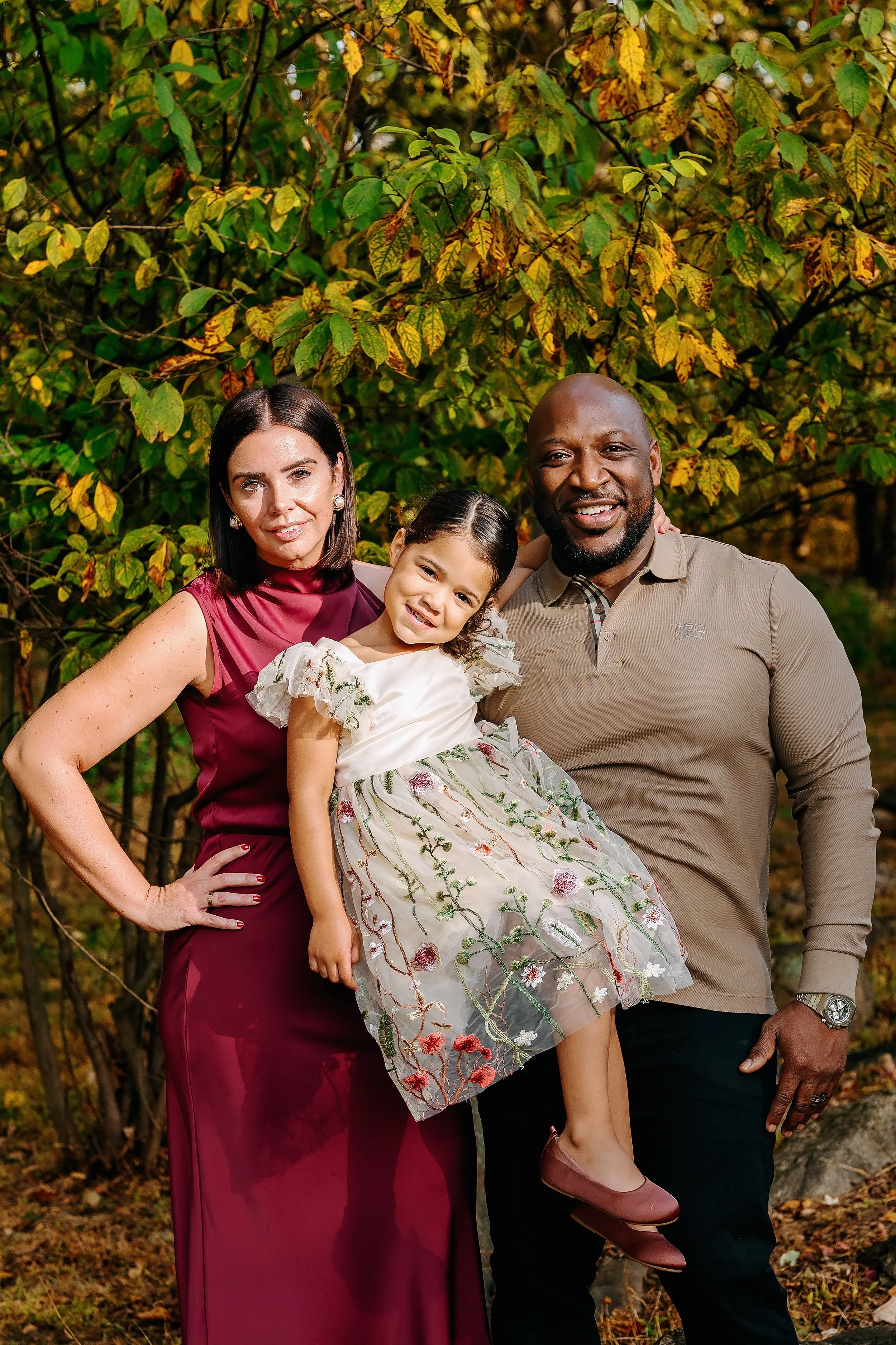 A family of three posing outdoors among autumn trees. The woman is wearing a sleeveless burgundy dress, the man is in a beige shirt, and the young girl is in a floral embroidered dress. The girl is sitting on the woman’s hip, and all are smiling.