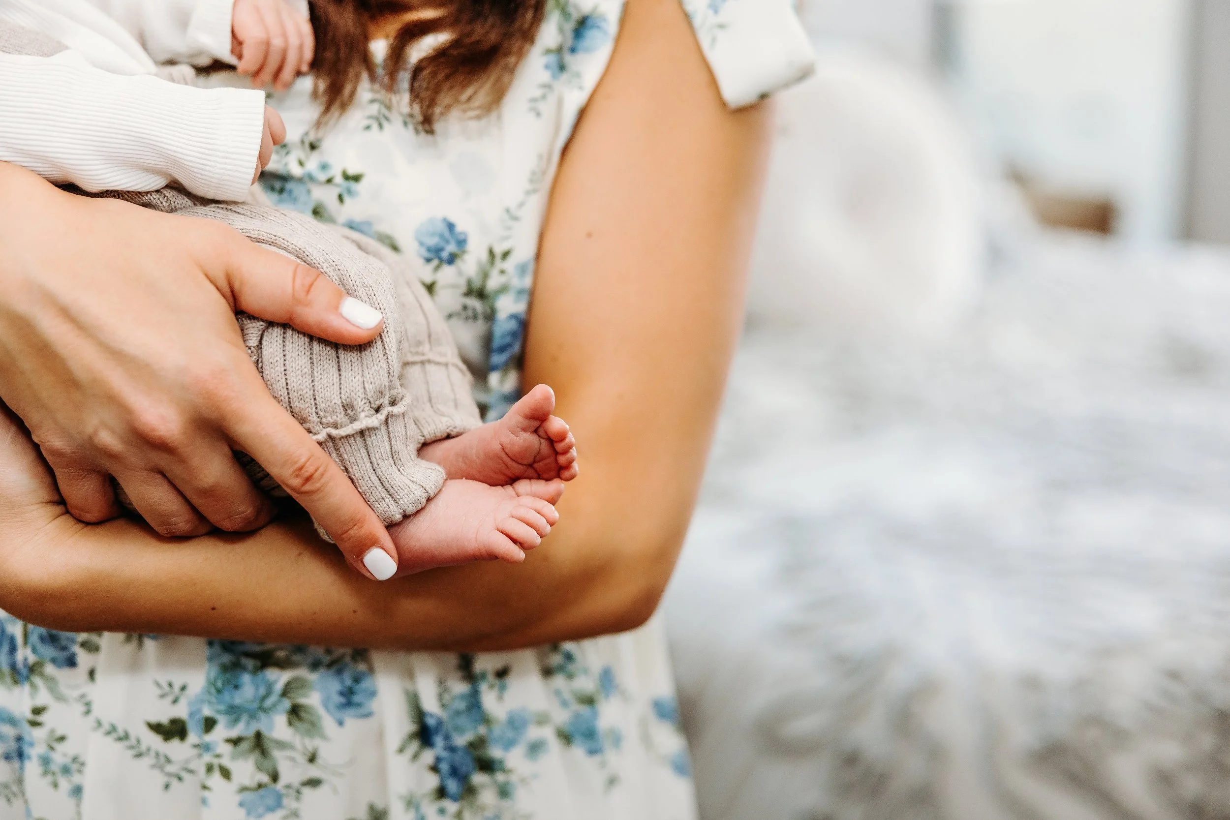 Close-up of a woman holding a tiny baby newborn. The woman wears a floral dress, and the baby is dressed in beige knit pants. The focus is on the baby's feet and the woman's hand supporting them.