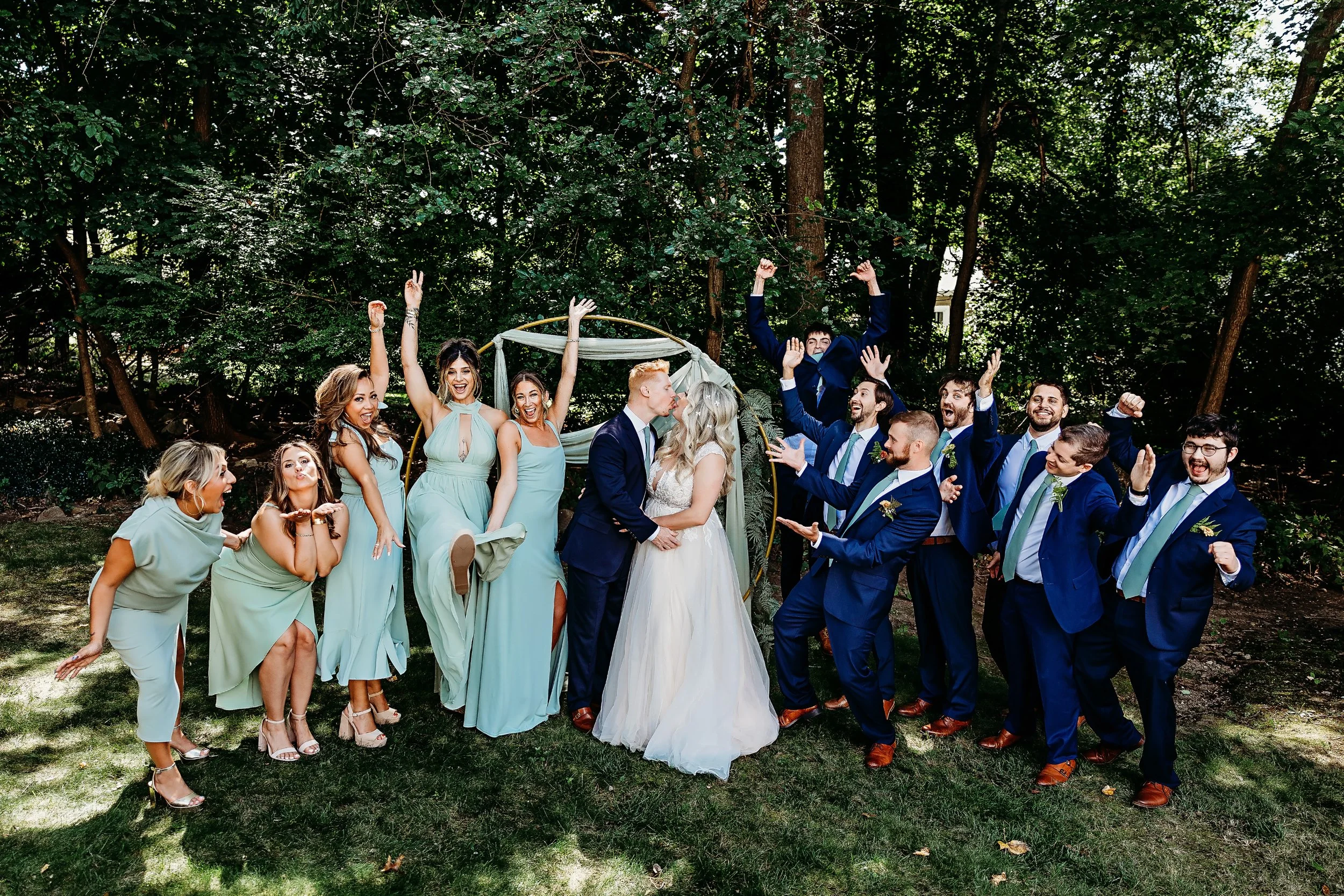 A wedding party celebrating outdoors with the bride and groom kissing and their friends cheering and posing joyfully.