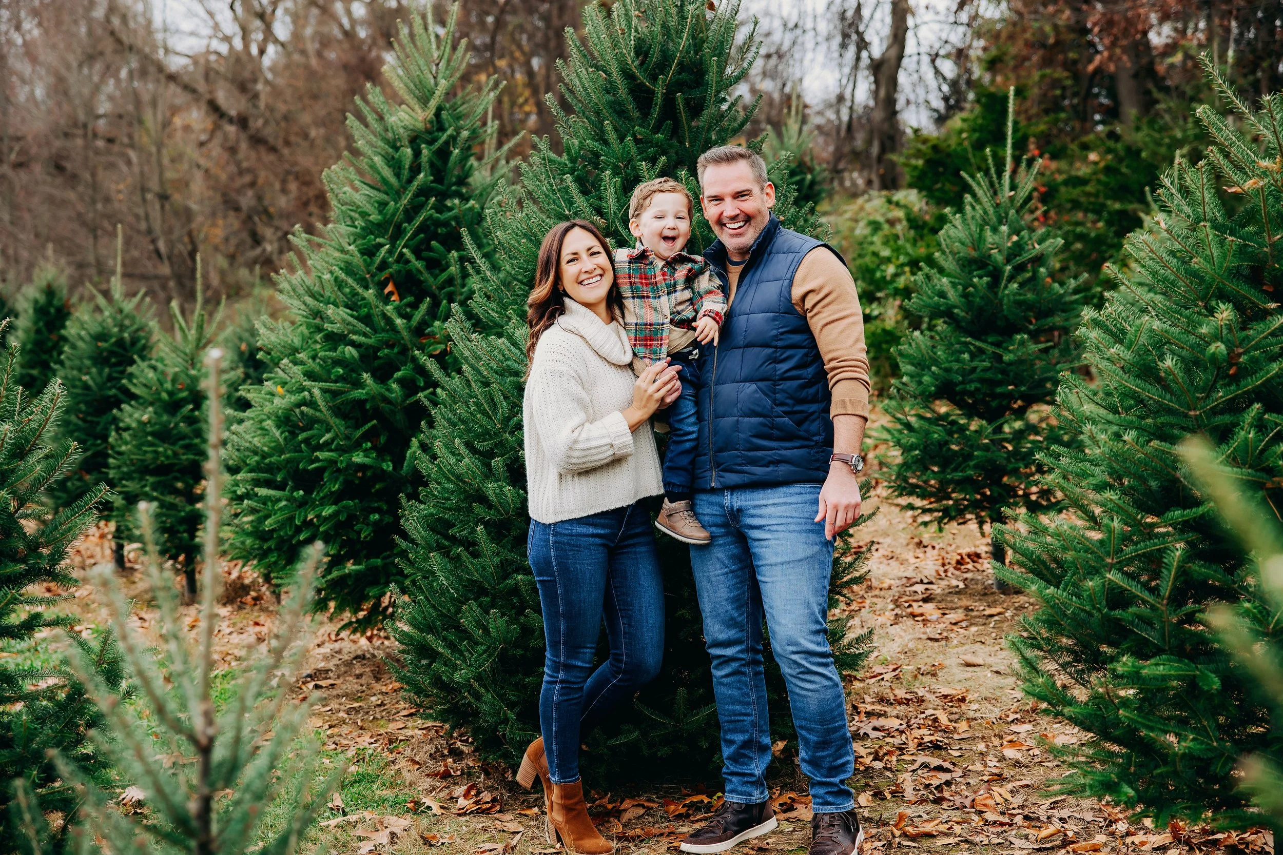 A happily smiling family of three standing among Christmas trees outdoors in autumn, with fallen leaves on the ground. The mother has long brown hair, is wearing a cream sweater and jeans. The father has short hair, is wearing a navy vest over a brown long-sleeve shirt and jeans. Their young son, with curly hair, is wearing a plaid shirt and khakis, and is being held by the mother.