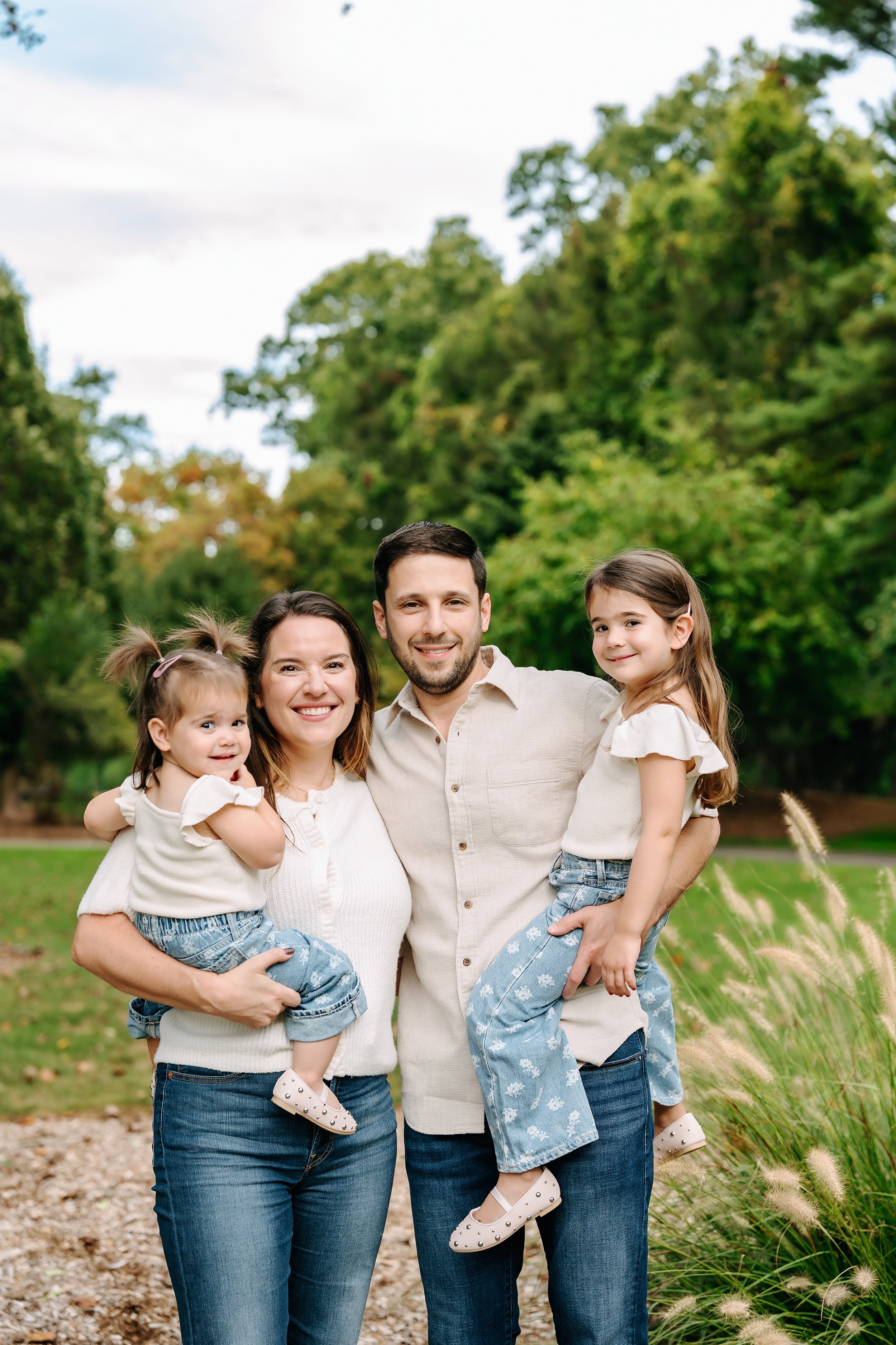 A family of four smiling and posing together outdoors in a park, surrounded by green trees and tall grass.