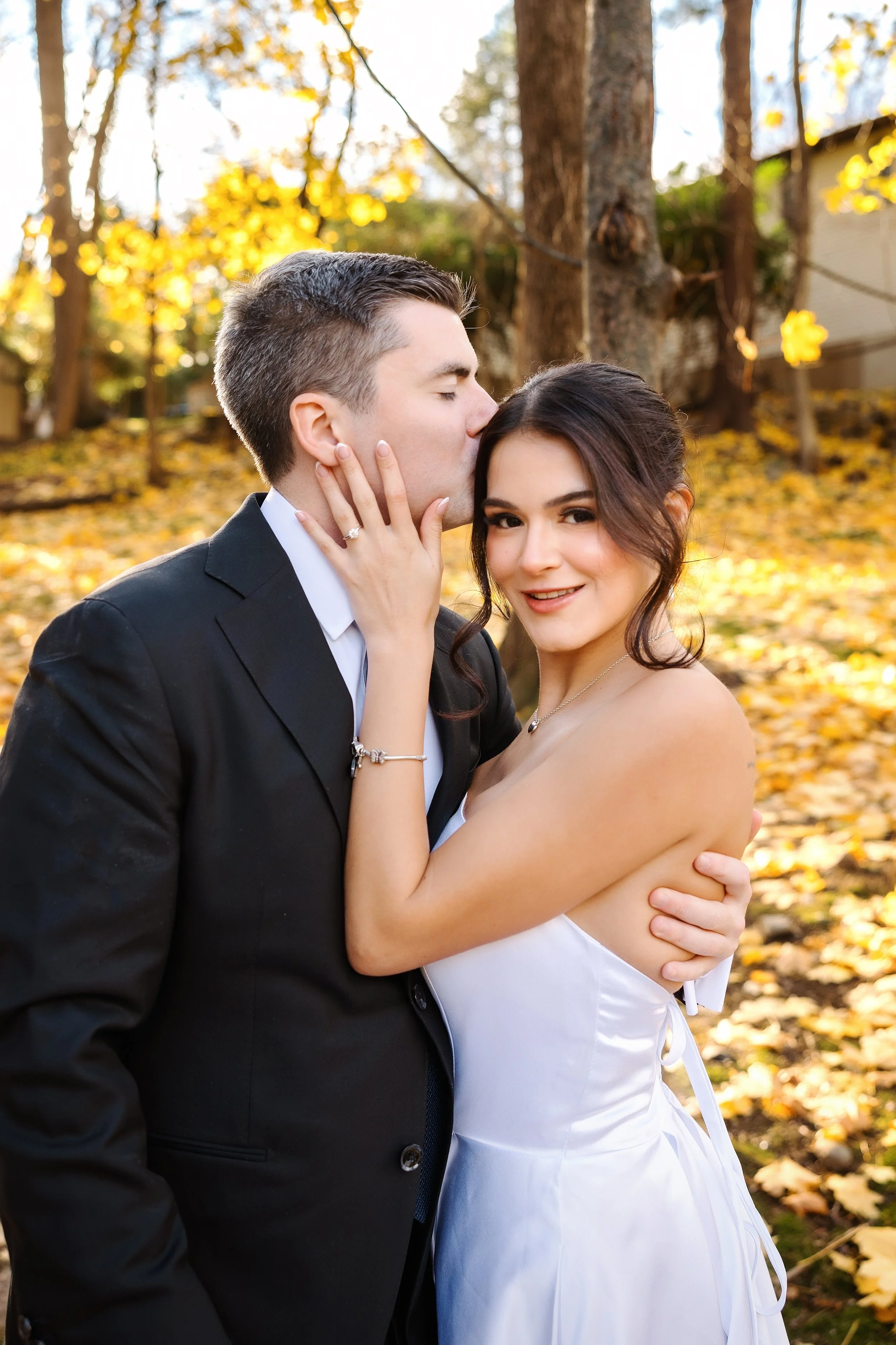 A young couple in formal attire embraces outdoors among fall foliage; the man is kissing the woman on the forehead, who is smiling and looking at the camera.