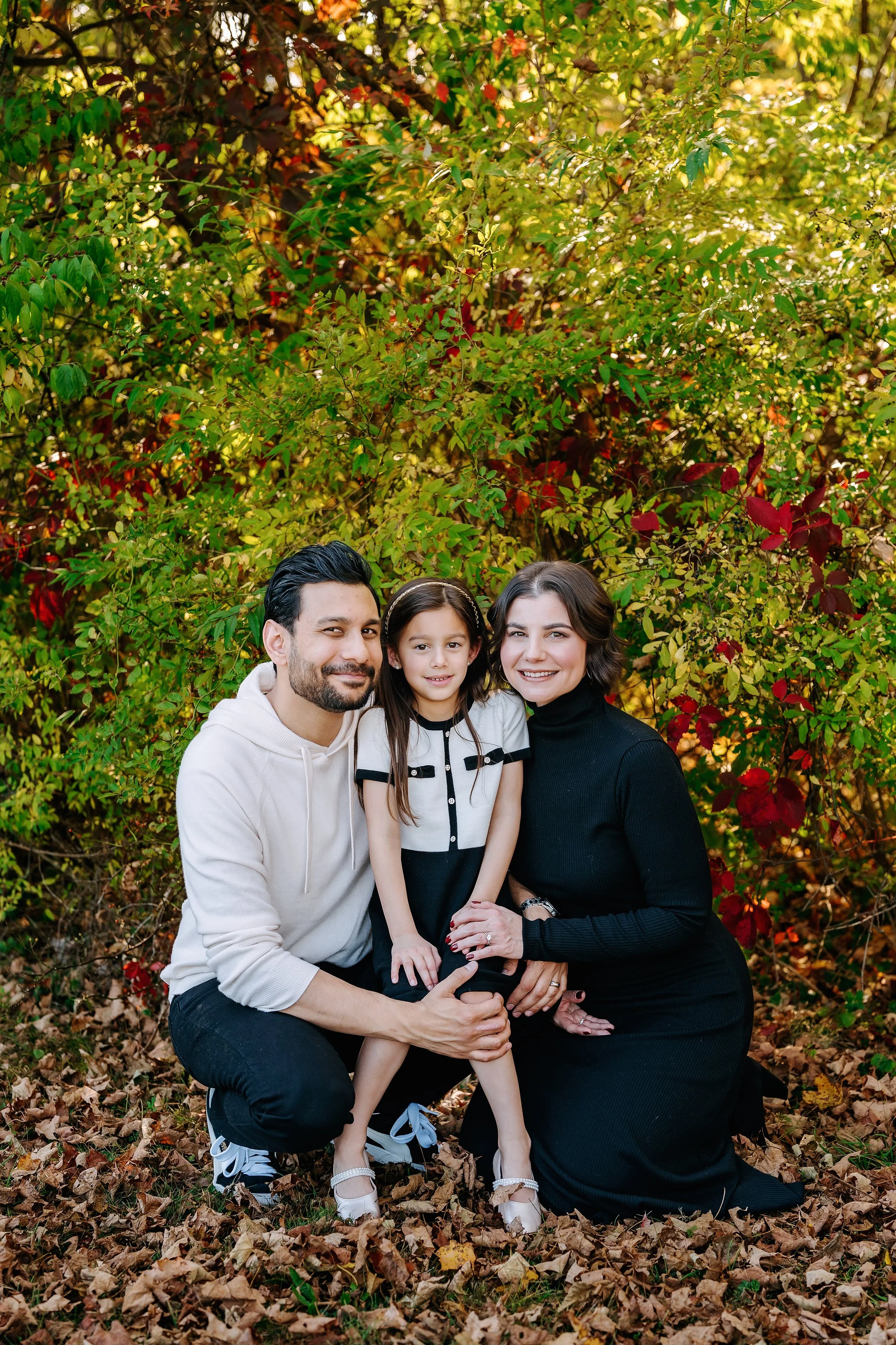 A family of three, a man, woman, and young girl, smiling outdoors surrounded by autumn foliage and fallen leaves.