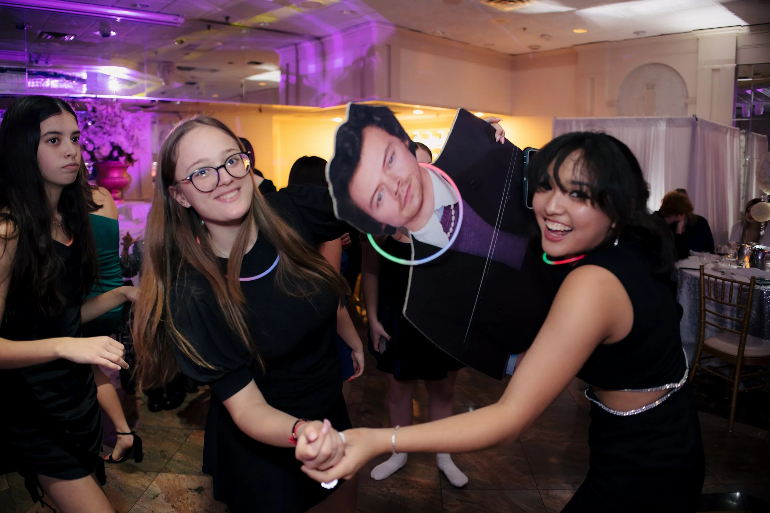 A group of young women dancing and celebrating at a party, with pink and purple lighting, some wearing glow necklaces, and one woman wearing a large walking pose sign around her neck.