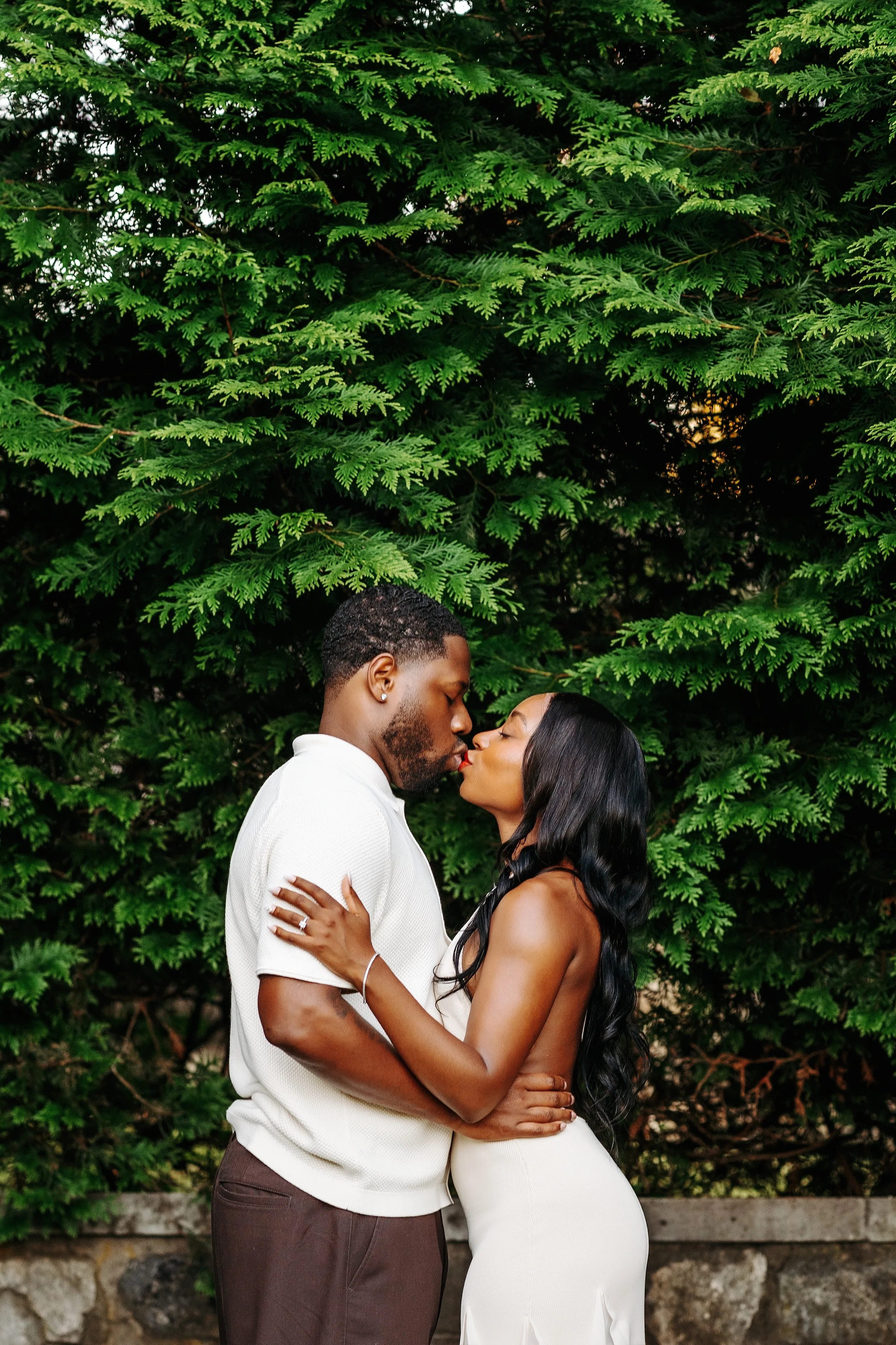 A couple kissing outdoors in front of lush green trees.