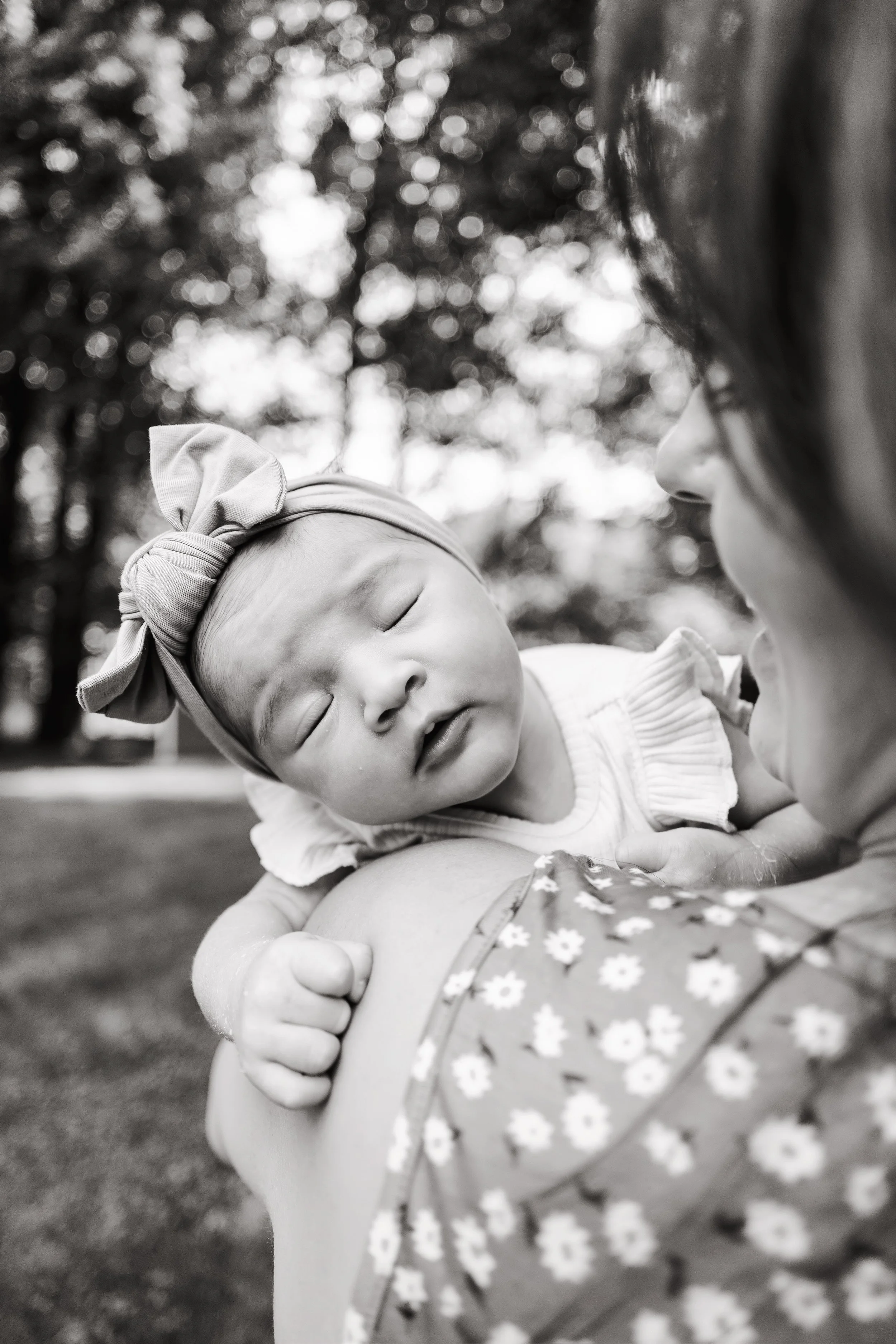 A black and white photo of a sleeping baby girl with a headband, resting on an adult's shoulder outdoors, with trees in the background.