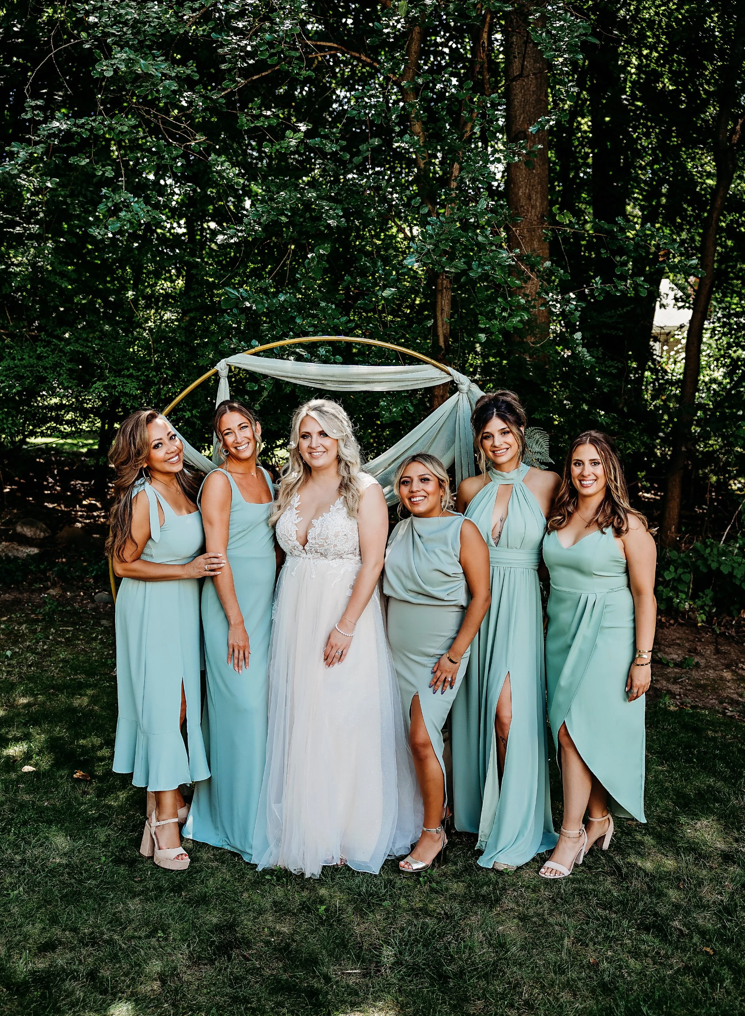 A bride in a white wedding dress standing with five bridesmaids in matching light blue dresses outdoors under trees.