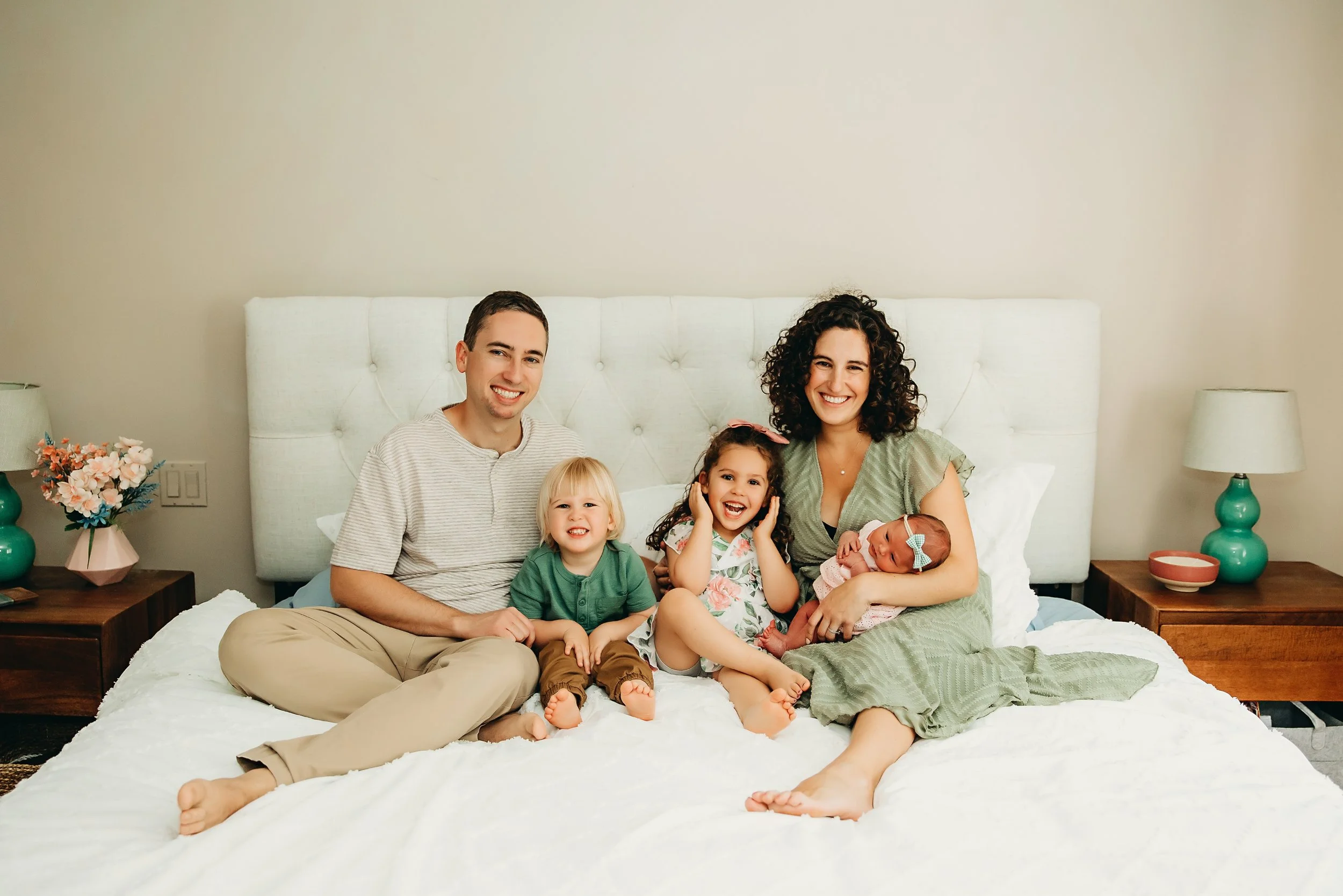 Family of five sitting on a bed in a neutral-colored bedroom, smiling, with a mother holding a newborn girl with a bow on her head, two young children sitting between parents, and a father in casual wear.